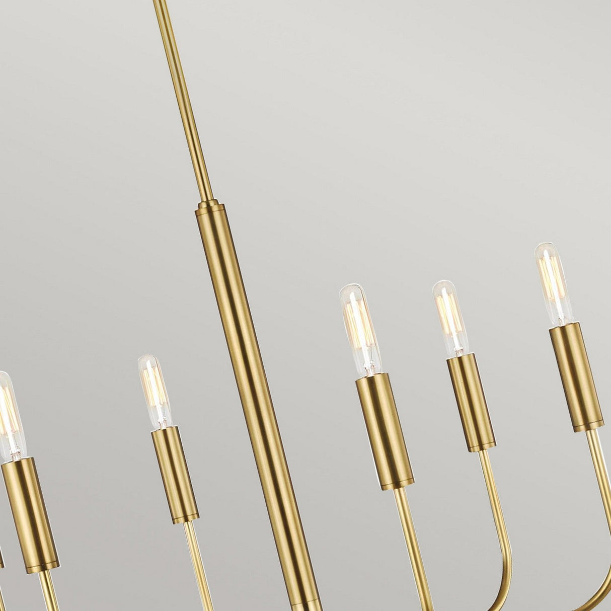The Brianna 6 Light Chandelier - Burnished Brass embodies modern elegance with its sleek, minimalist design. With several vertical rods crowned with exposed bulbs, it creates a striking contrast against a light gray backdrop.