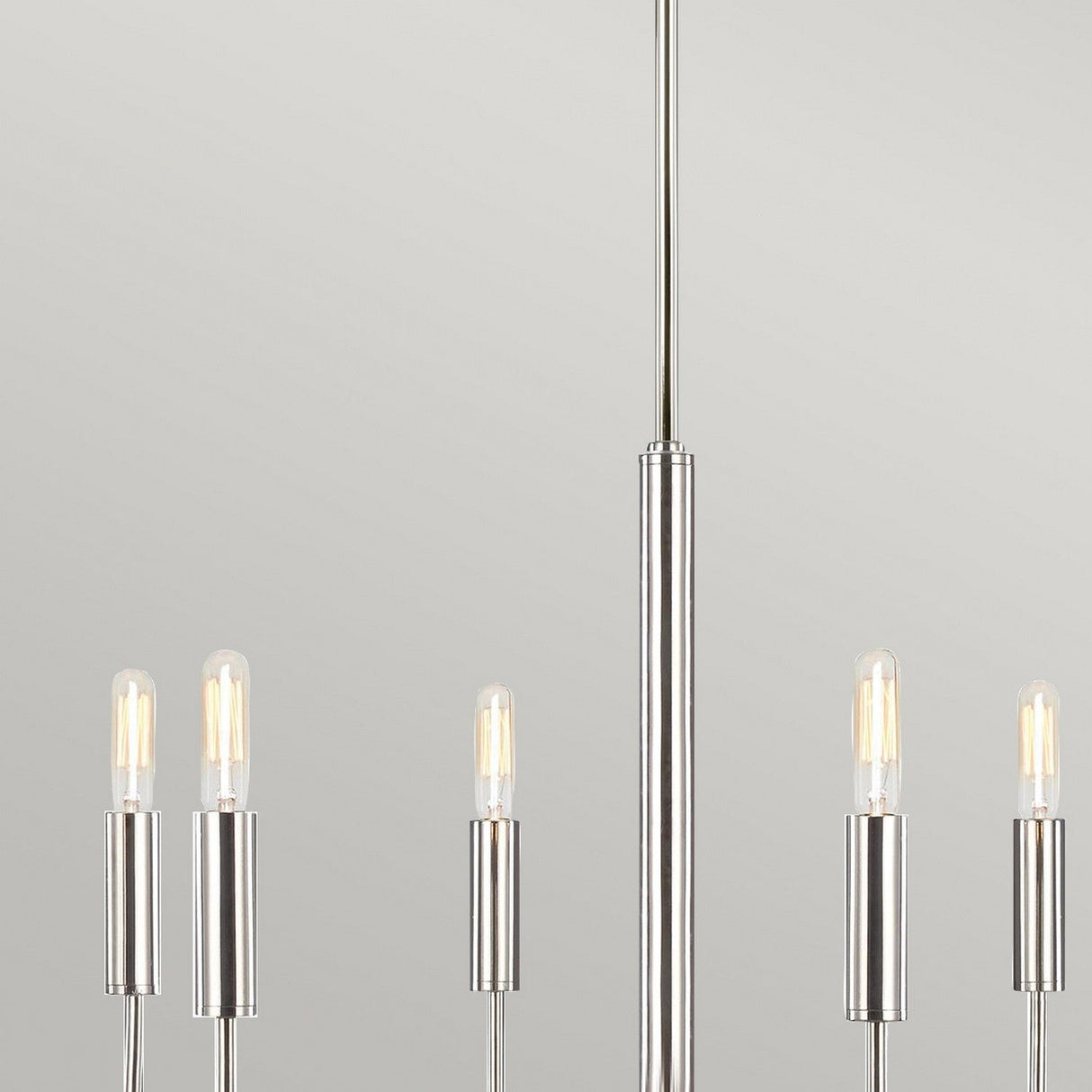 The Brianna 6 Light Chandelier - Polished Nickel adds a touch of elegance to modern interiors, boasting a sleek metal structure. It features six exposed bulbs in cylindrical holders arranged around a vertical rod against a light gray background.