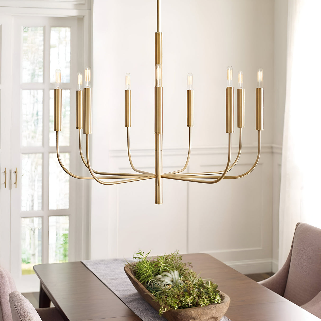 A contemporary dining room features an elegant design with the Brianna 9 Light Chandelier - Burnished Brass, casting a warm glow above a wooden table. The table is embellished with a grey runner and lively greenery, as gentle natural light streams through sheer curtains.