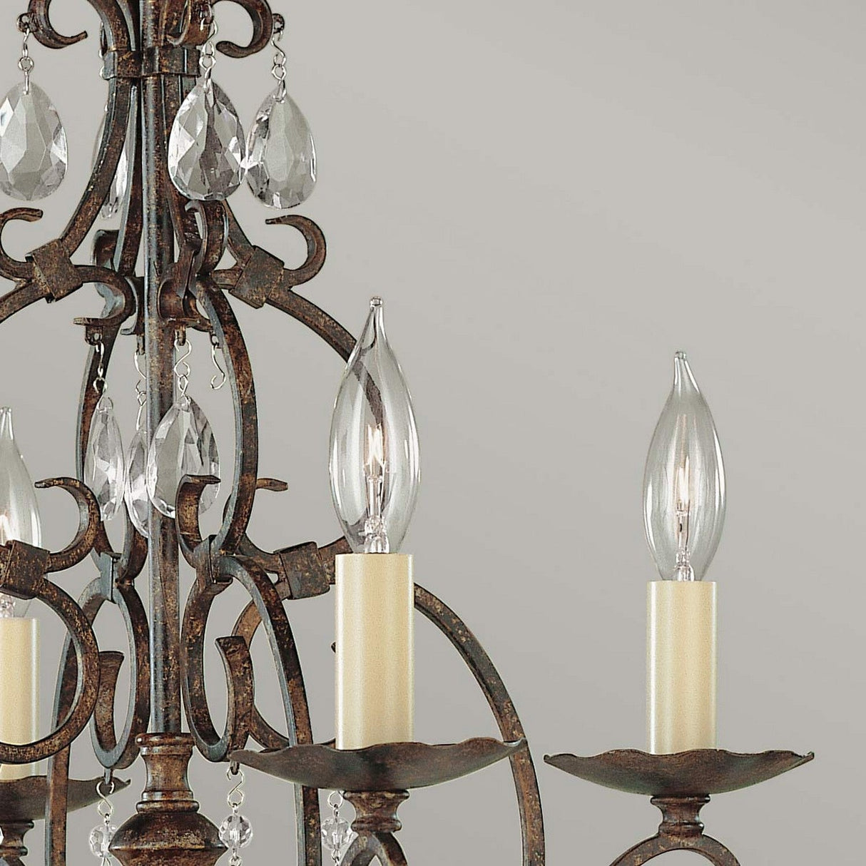 A detailed view of the Chateau 4 Light Chandelier reveals its ornate design with decorative crystal accents and candle-shaped light bulbs on a gracefully curved metal frame, set against a neutral backdrop. The metal is finished in mocha bronze, enhancing its rustic elegance.