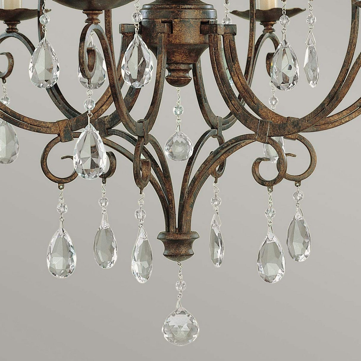 A detailed look at the Chateau 6 Light Chandelier - Mocha Bronze, which is embellished with teardrop-shaped crystal accents. The decorative metal frame displays elaborate scrollwork, with crystals gracefully hanging and reflecting light against a subtle, neutral gray backdrop.