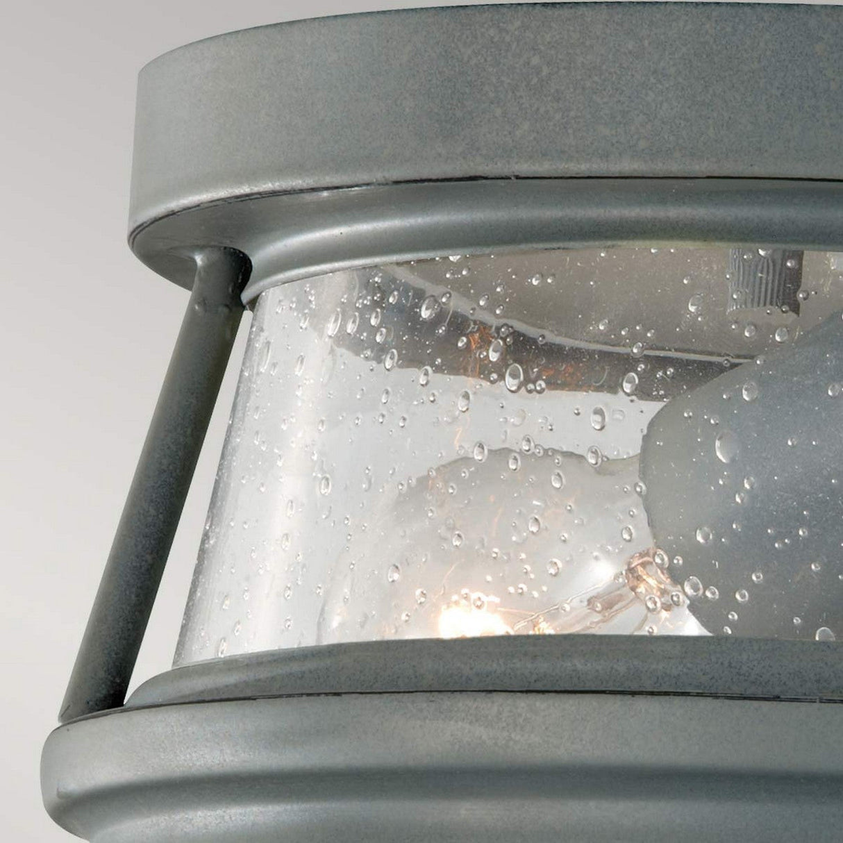Close-up of a Chelsea Harbor 2 Light Outdoor Flush Light - Storm Cloud, showcasing its clear glass panel adorned with water droplets. This coastal design features a warm glow emanating from within its cylindrical shape, framed in metal for durability.