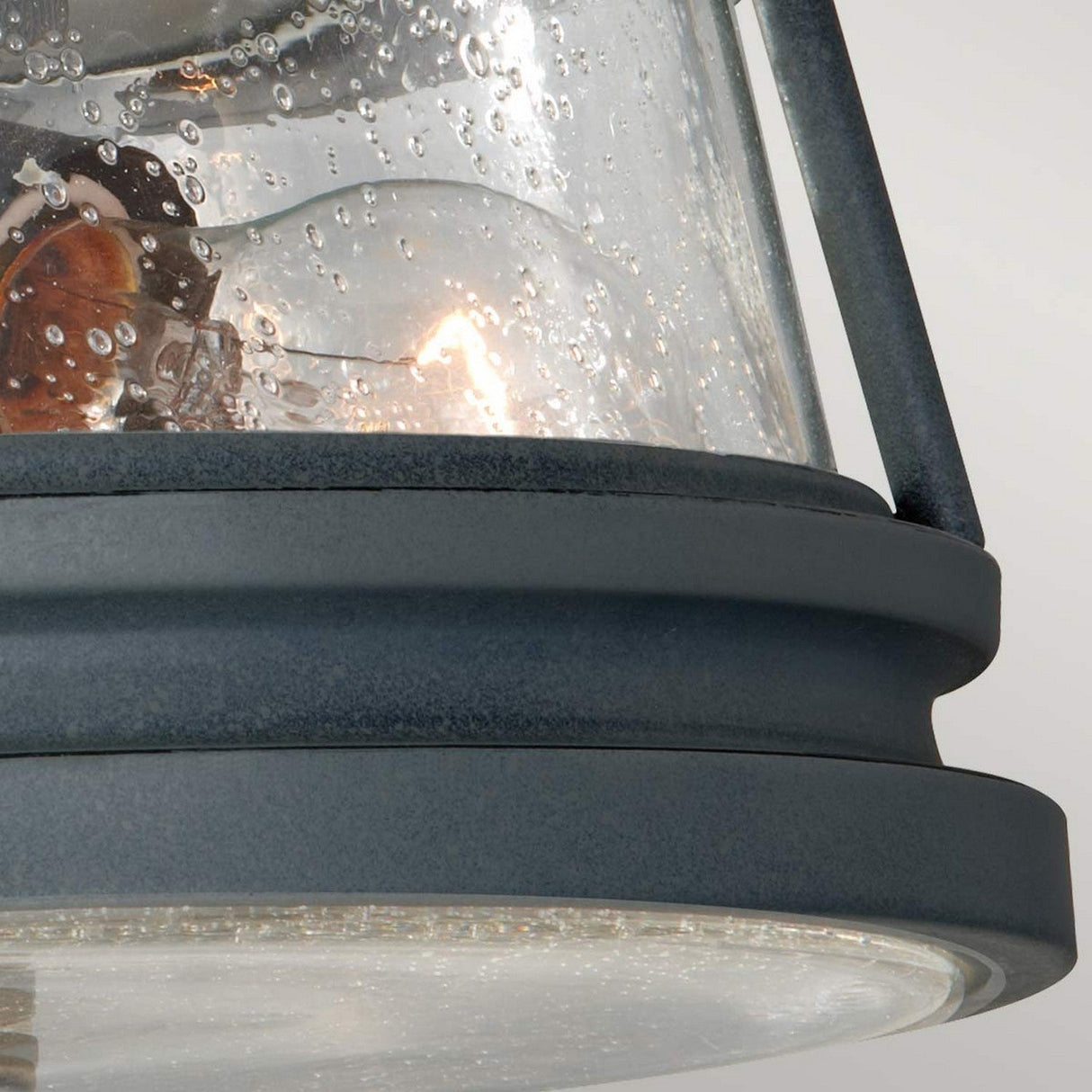 A close-up of the Chelsea Harbor 2 Light Outdoor Flush Light - Storm Cloud shows its modern design with a clear, textured glass cover adorned with small bubbles, through which a glowing bulb is visible. The dark, matte frame accentuates its contemporary coastal aesthetic.