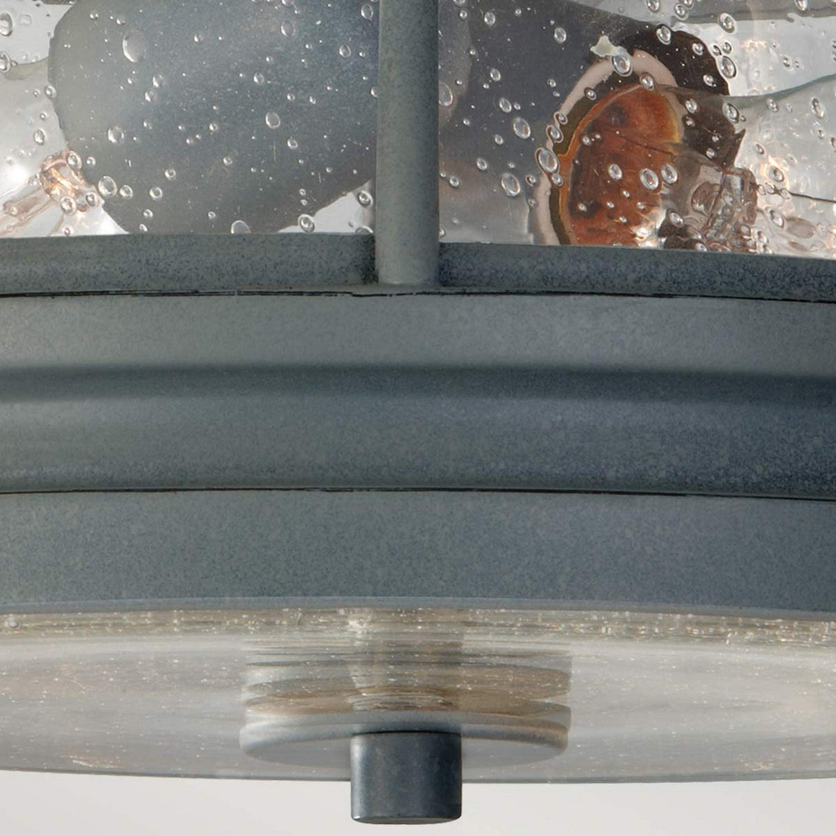 Close-up of the Chelsea Harbor 2 Light Outdoor Flush Light - Storm Cloud, featuring a black metal design with clear, textured glass casing. Reminiscent of coastal style, the fixture is adorned with dewy water droplets. The inner bulb is partially visible through the glass, evoking timeless elegance.