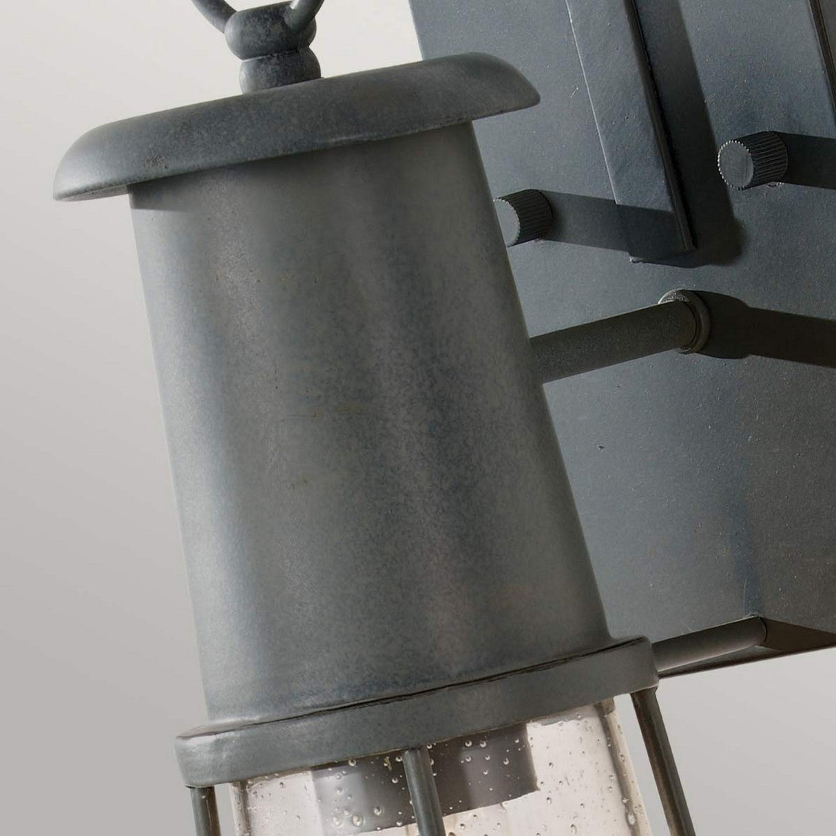 Close-up of the Chelsea Harbor 1 Light Outdoor Wall Lantern in a vintage aesthetic and rustic style. This fixture showcases a cylindrical top and clear glass shade speckled with visible bubbles, all mounted on a metal arm with a storm cloud finish. The background features a plain light color.