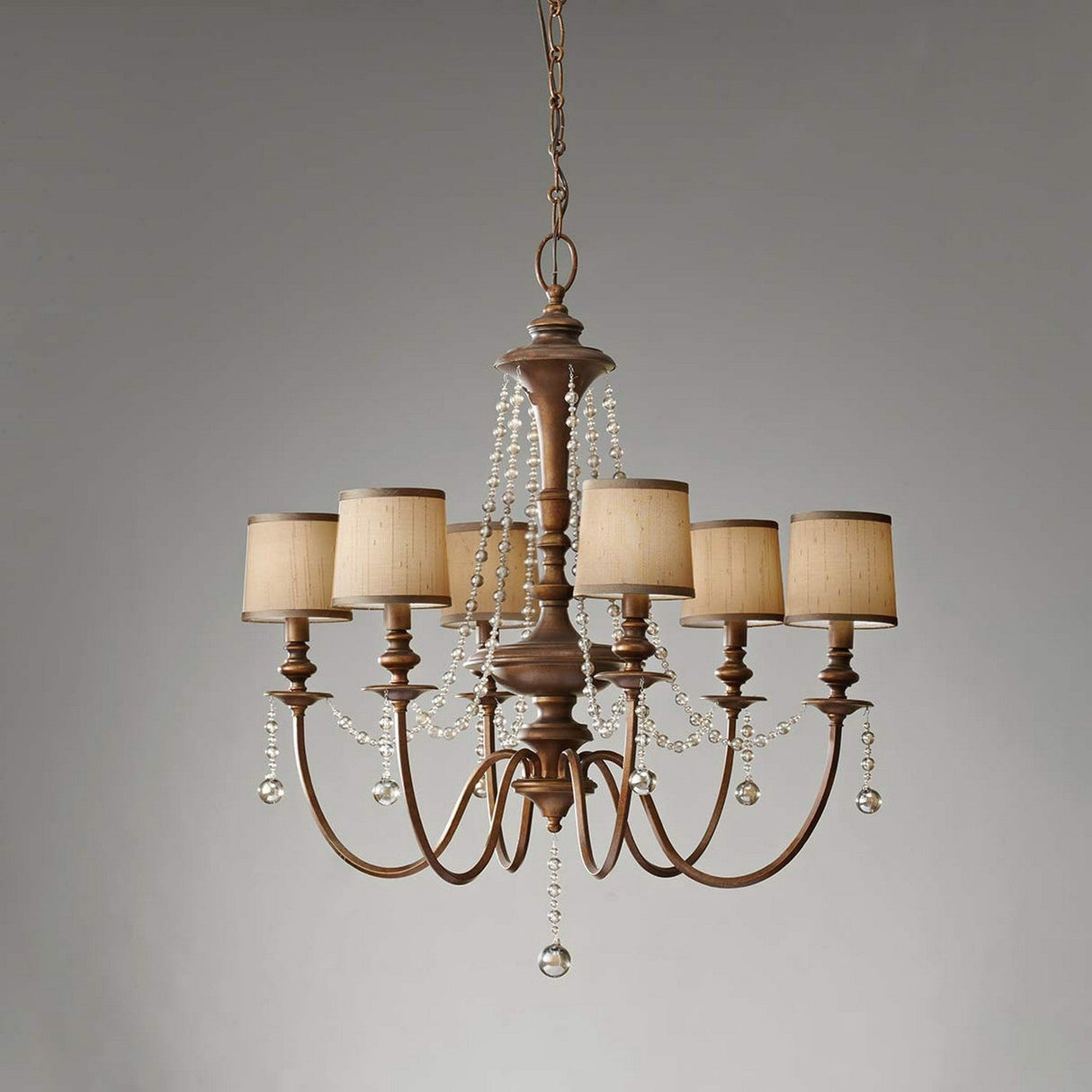 The Feiss Clarissa 6 Light Chandelier in a Firenze Gold finish showcases a vintage style with its six arms and beige lampshades. It includes decorative chains of champagne-plated glass beads that elegantly hang between the arms, creating a sophisticated contrast against a neutral backdrop.