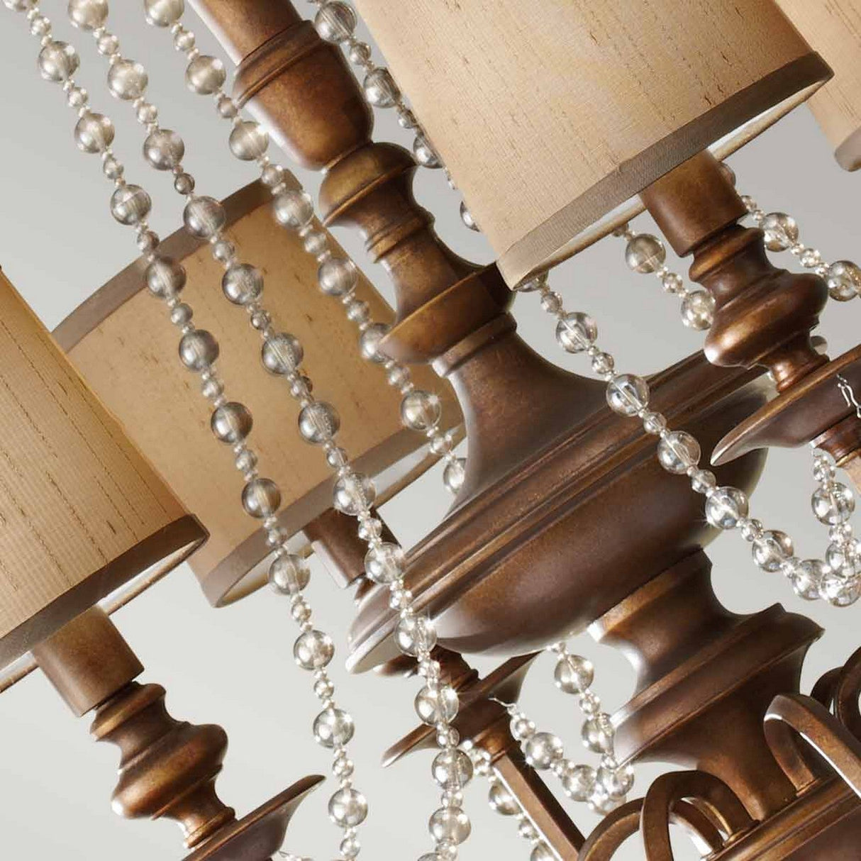 The Feiss Clarissa 6 Light Chandelier, finished in Firenze Gold, showcases a detailed bronze design with multiple light fixtures adorned with beige drum-shaped shades. Its structure is draped with champagne-plated glass beads, enhancing the chandelier's sophisticated elegance.