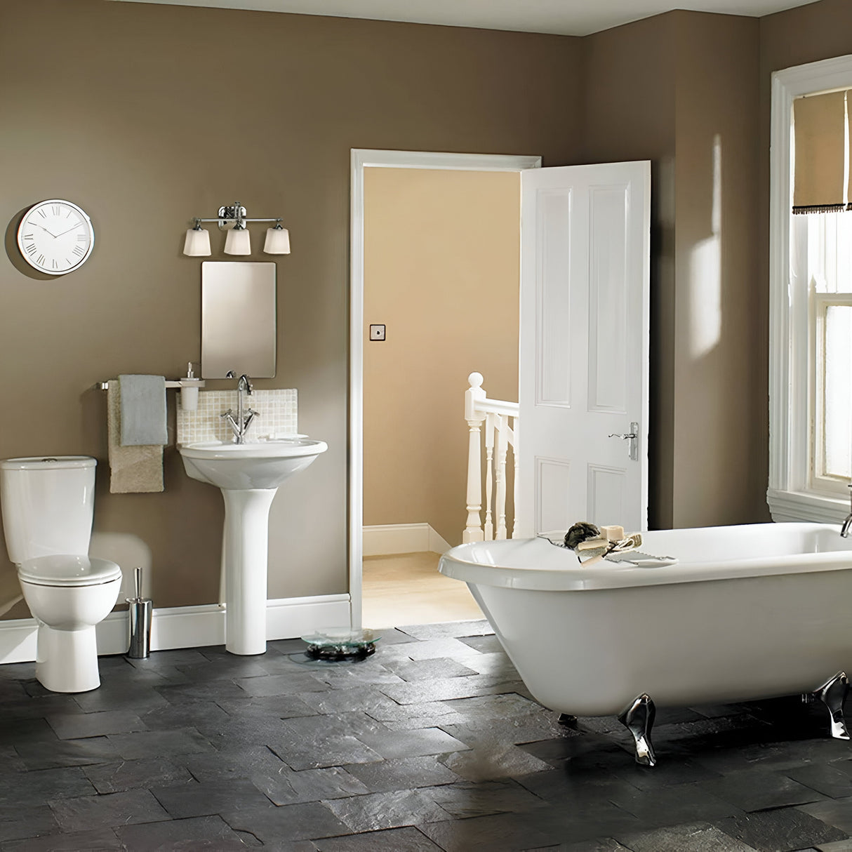 A modern bathroom showcasing a clawfoot bathtub, pedestal sink, and toilet is adorned with polished chrome accents. The room's neutral color palette is brightened by the Concord 3 Light Wall Light in Polished Chrome with opal-etched glass shades, while sunlight filters through a window partially covered by a blind.