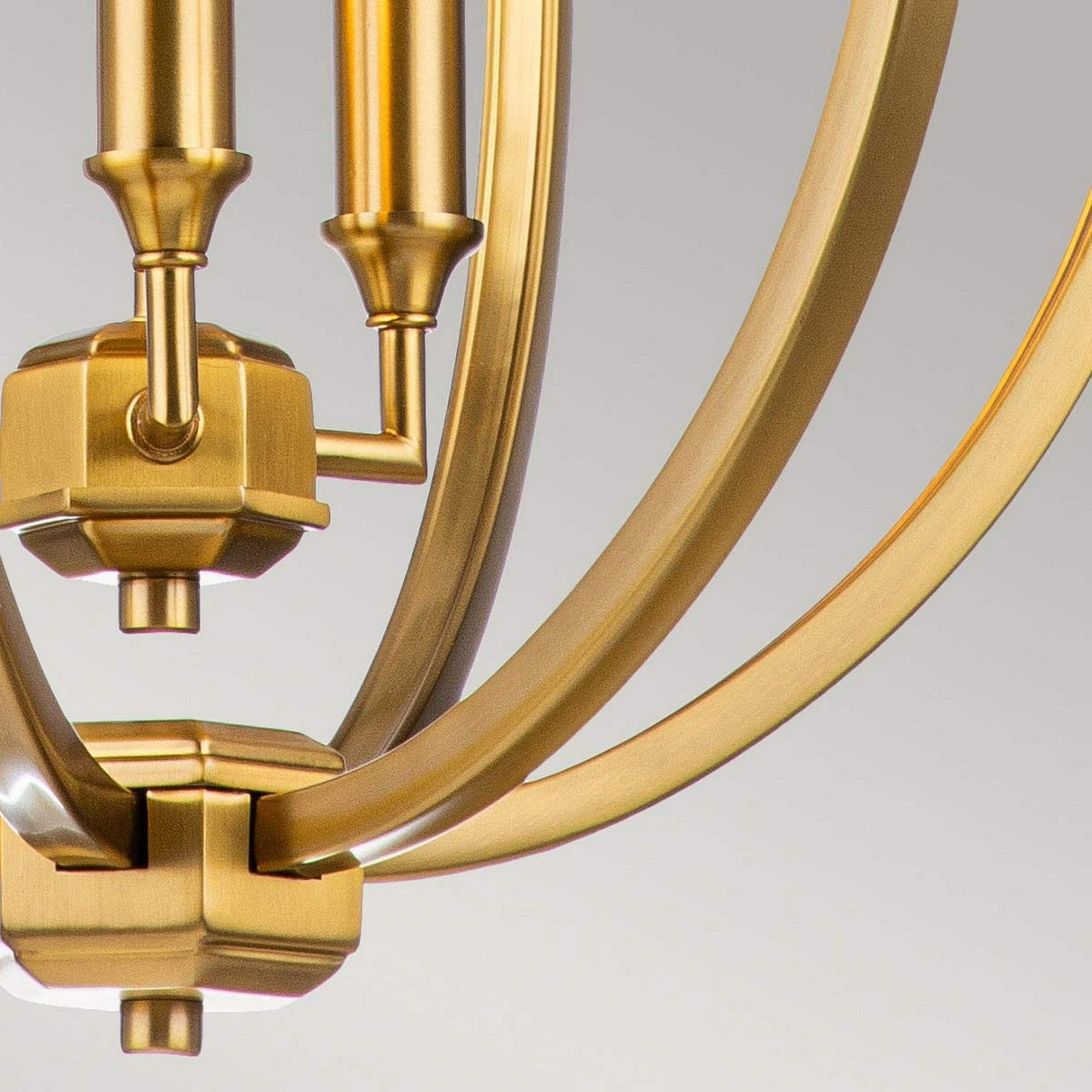 The Corinne 3 Light Medium Chandelier, made from burnished brass, features a geometric design with curved metal lines and small cylindrical elements. Its modern aesthetic is accentuated by subtle mid-century design influences, beautifully highlighted against a neutral background in this stunning close-up view.