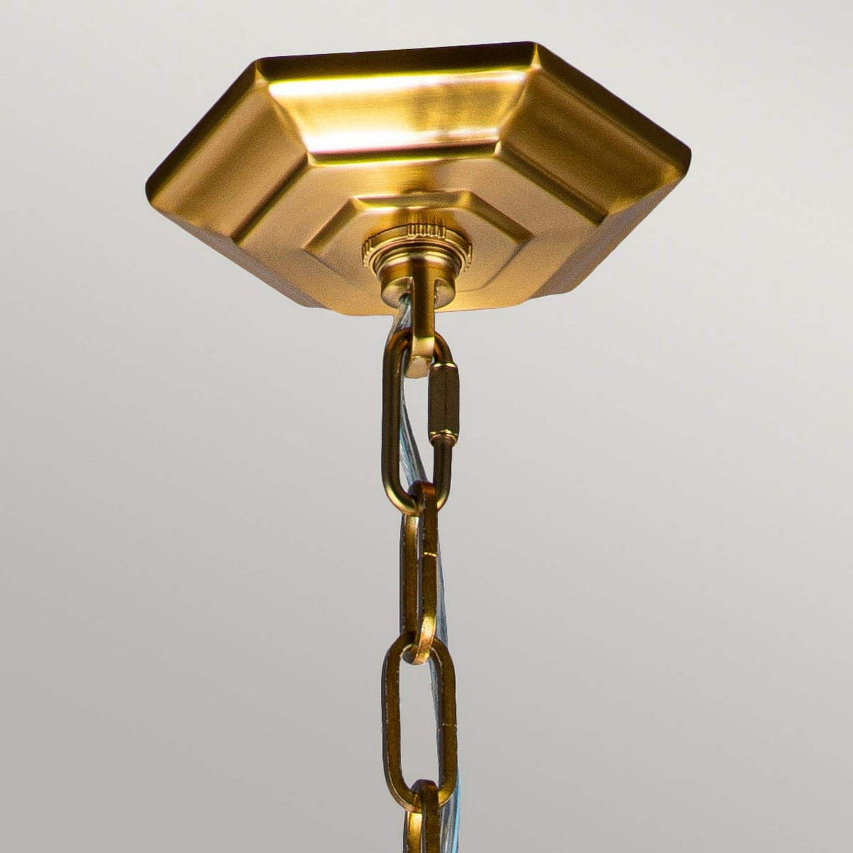 The Corinne 3 Light Medium Chandelier in burnished brass offers a close-up view of its hexagonal design, featuring a metal chain that suspends from the fixture. The sturdy links of the chain highlight the chandelier's Mid-Century geometric elegance against a simple, light-colored background.