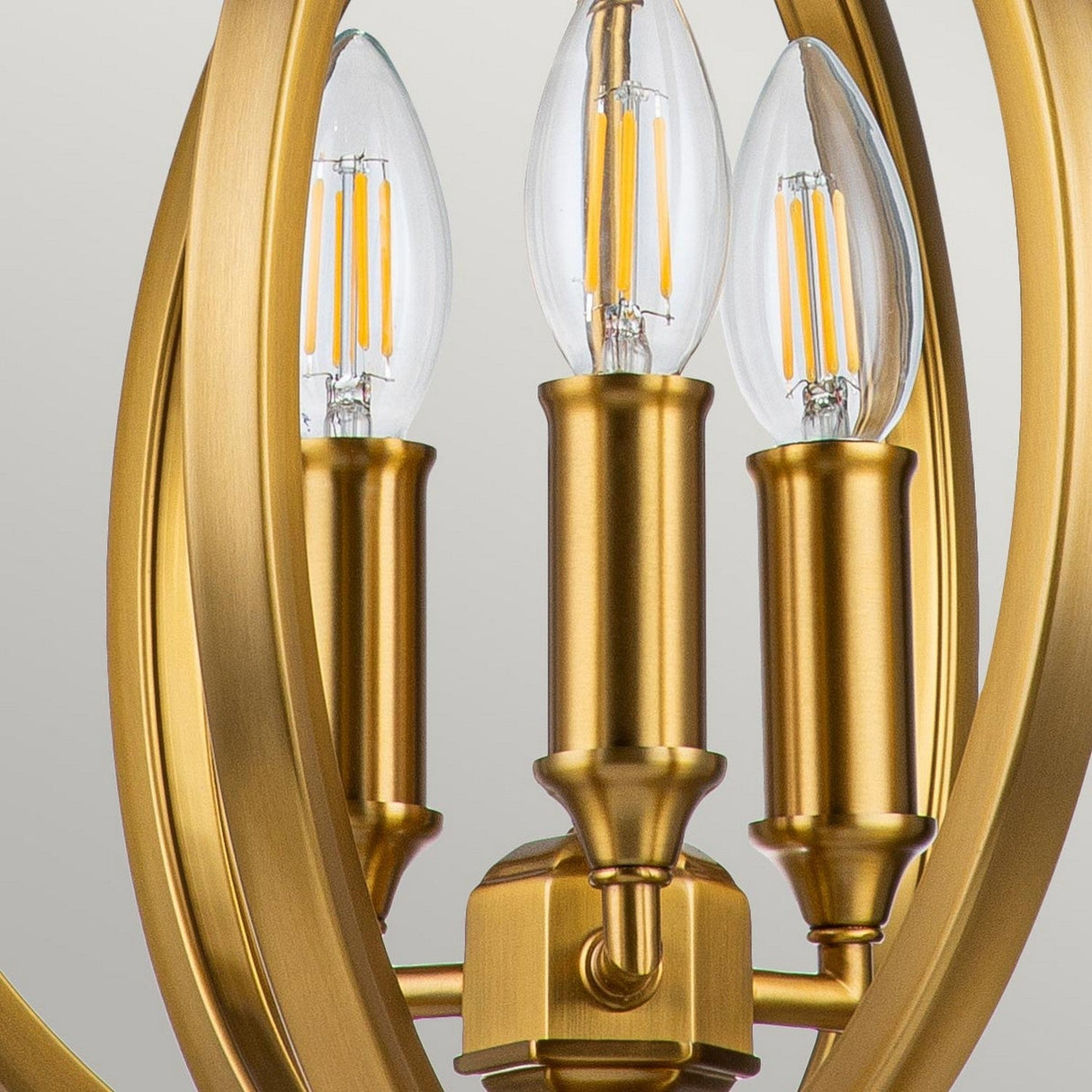 A close-up of the Corinne 3 Light Small Chandelier in burnished brass reveals its three candle-style LED bulbs framed by a circular structure. This elegant lighting fixture boasts a modern design, exuding sophistication and charm.