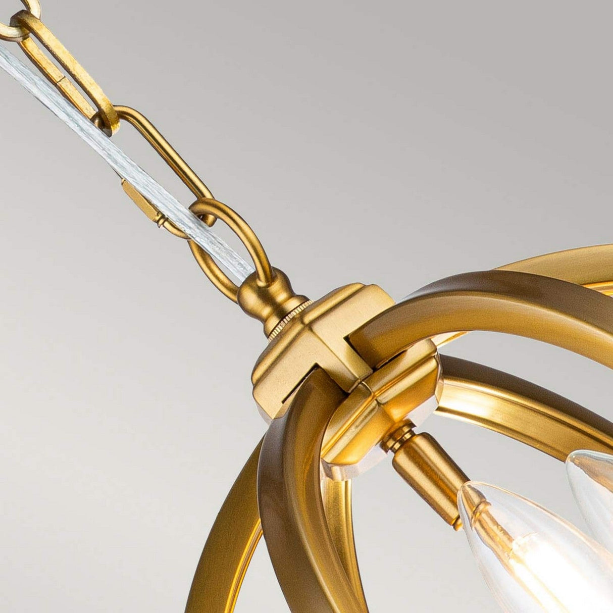 A close-up view showcases the Corinne 3 Light Small Chandelier in Burnished Brass, revealing its modern gold aesthetic with a decorative circular design. This elegant fixture features exposed bulbs and hangs gracefully from a burnished brass chain against a plain background.