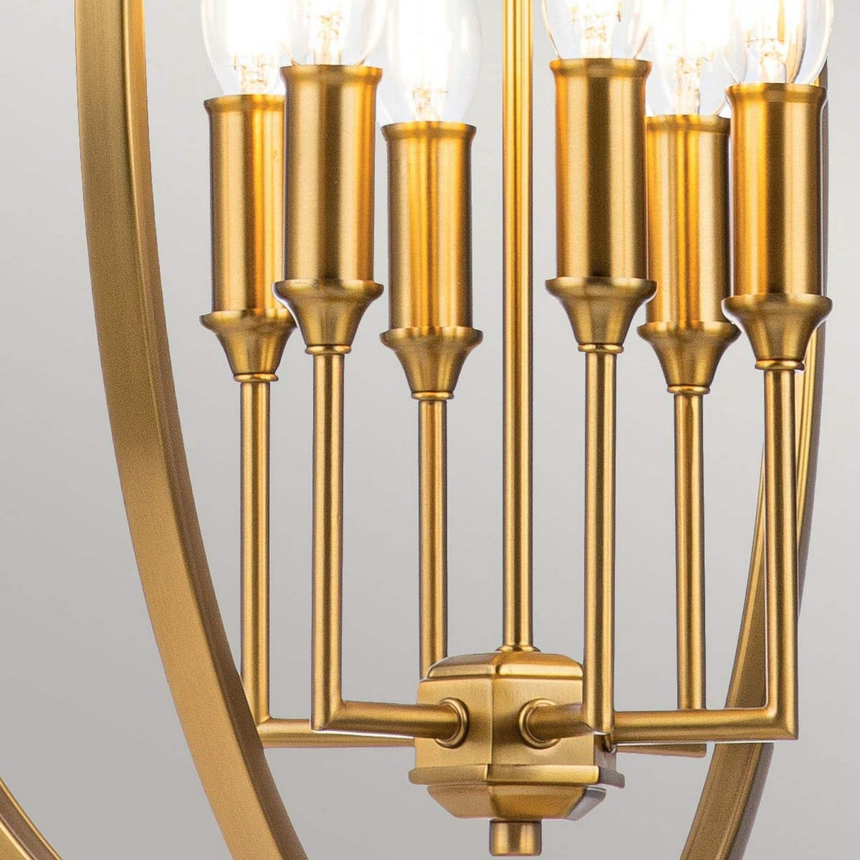 Close-up of the Corinne 6 Light Chandelier in burnished brass featuring exposed clear bulbs. Its design showcases sleek cylindrical holders and a series of vertical rods, forming an elegant open orb silhouette against a neutral background.