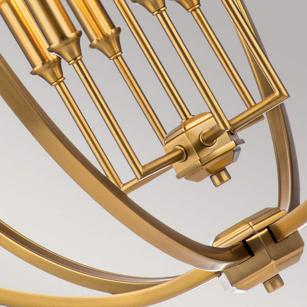 The Corinne 6 Light Chandelier in Burnished Brass is highlighted in a close-up, revealing its mechanical structure with gears and parallel rods. The intricate design of this chandelier showcases metallic finishes and cylindrical elements arranged in a curved framework, evoking the silhouette of an open orb against a light background.