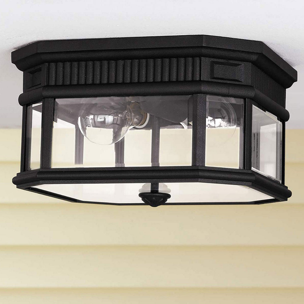 The Cotswold Lane 2 Light Outdoor Flush - Black is an octagonal ceiling light fixture with bevelled glass panes and two prominent light bulbs. This sophisticated black lantern fixture is mounted on a white ceiling, complemented by a beige wall in the background, enhancing the elegance of any space.