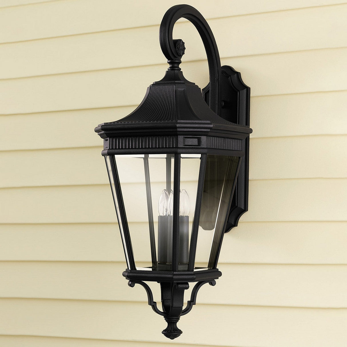 A Cotswold Lane 3 Light Outdoor Large Wall Lantern in a black finish, featuring clear glass panels, is elegantly mounted on a cream-colored wall. This lantern, with its curved top and three candle-like bulbs inside, adds timeless charm to any exterior.