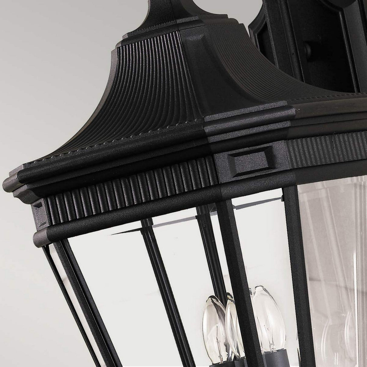 Close-up of the Cotswold Lane 3 Light Outdoor Large Wall Lantern in a black finish, exhibiting a classic design with clear glass panels and visible light bulbs inside. The detailed top section features vertical ridges and a decorative pattern.