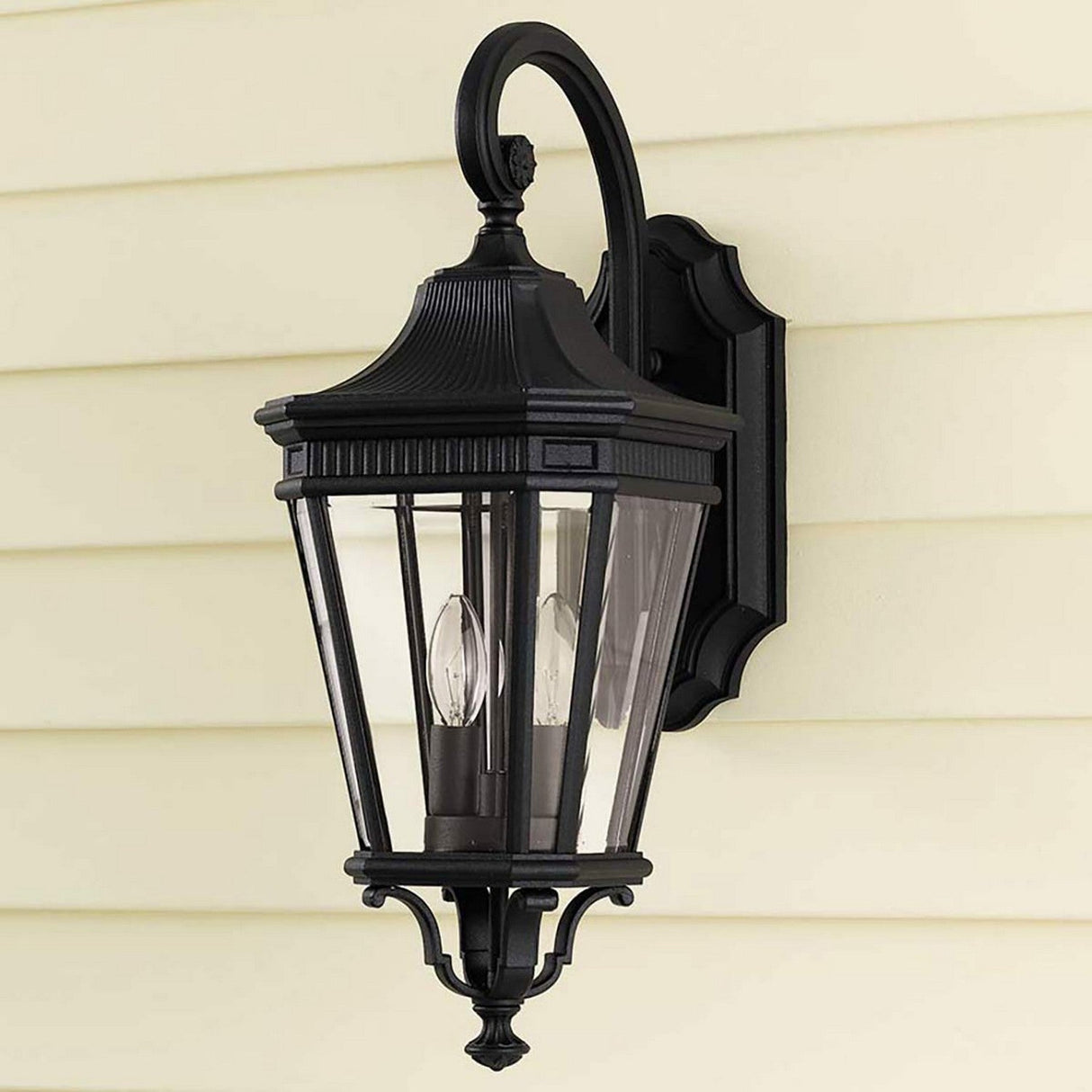 The Cotswold Lane 2 Light Outdoor Medium Wall Lantern in Black is a vintage-style wall light with clear glass panels and an elegantly curved arm. Mounted on a beige wall, it features two candle-shaped bulbs that illuminate your exterior with timeless charm.