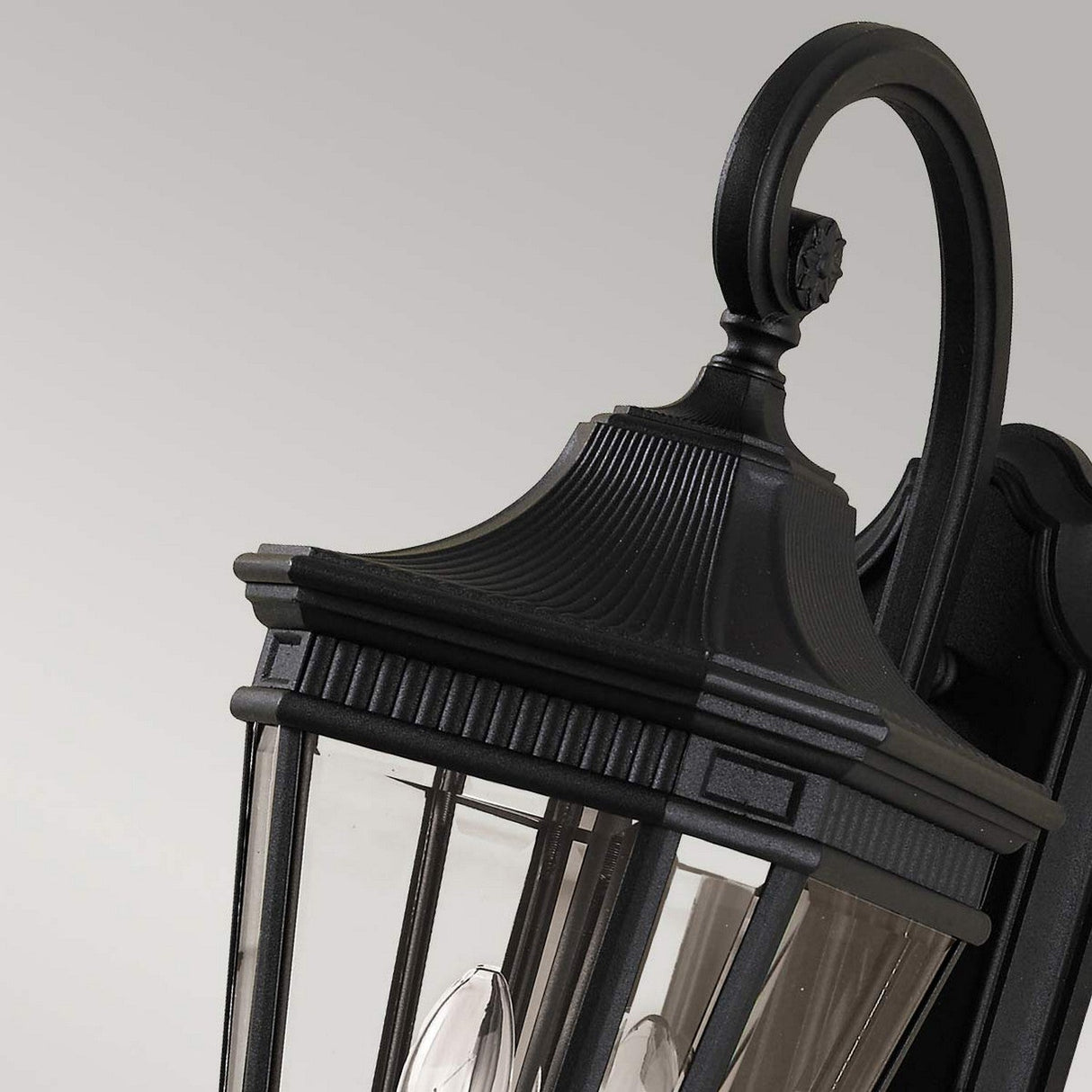 Close-up of the Cotswold Lane 2 Light Outdoor Medium Wall Lantern in black, featuring a decorative top and clear glass panels. The bulb filaments inside are beautifully showcased against a neutral gray background.