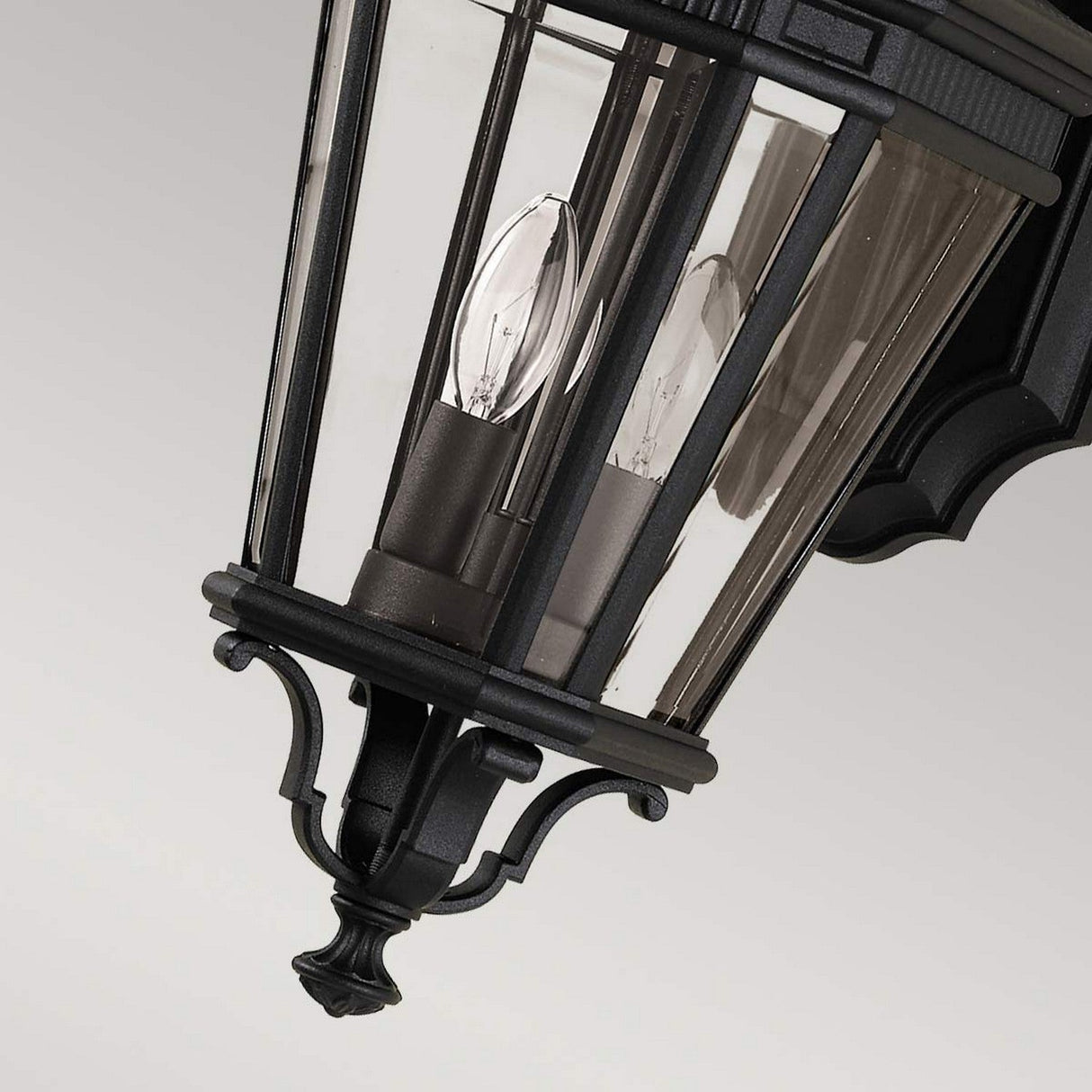The Cotswold Lane 2 Light Outdoor Medium Wall Lantern in black showcases clear glass panels with two candle-style bulbs, while ornate detailing at the base and top enhances its elegant design, making it a charming addition to any exterior space.