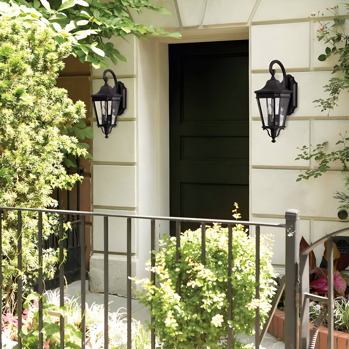 A charming entrance highlights a dark green door, framed by two Cotswold Lane 2 Light Outdoor Medium Wall Lanterns in Black. Cream-colored walls are decorated with greenery and potted plants, complemented by a Cotswold Lane style black metal fence that borders the front, enhancing the welcoming ambiance.