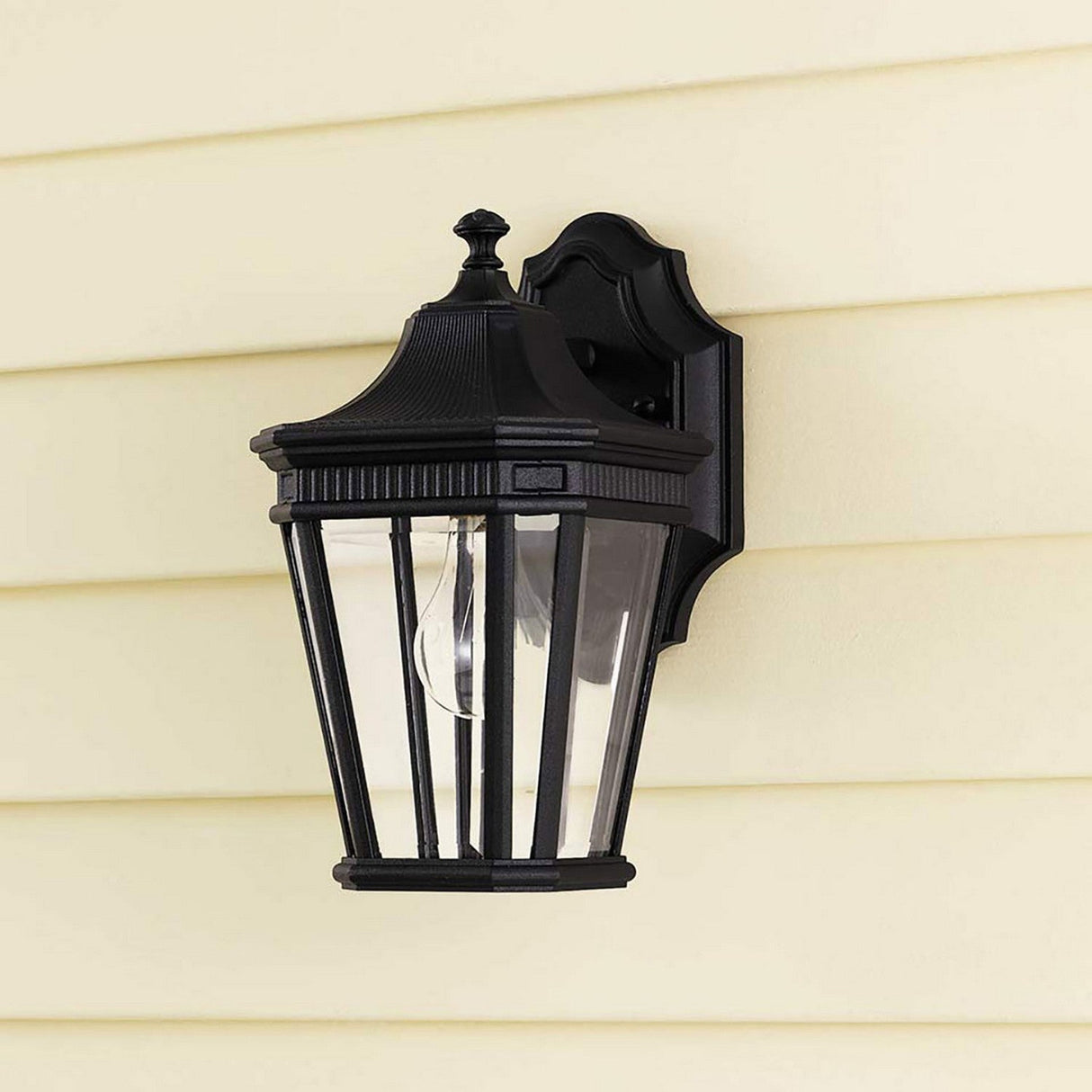 A Cotswold Lane 1 Light Outdoor Small Wall Lantern in a black finish is mounted on a light yellow siding wall. This vintage-style outdoor wall light features a clear glass cover with a visible bulb, adorned with decorative top and bottom trim and vertical bars surrounding the glass.