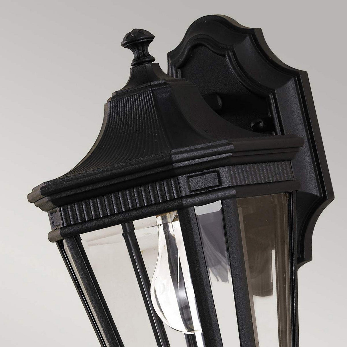The Cotswold Lane 1 Light Outdoor Small Wall Lantern in black boasts a traditional design with clear glass panels. It features a decorative top, vertical lines on the frame, and is mounted on a dark, shaped backplate. The visible clear bulb inside enhances its classic charm.