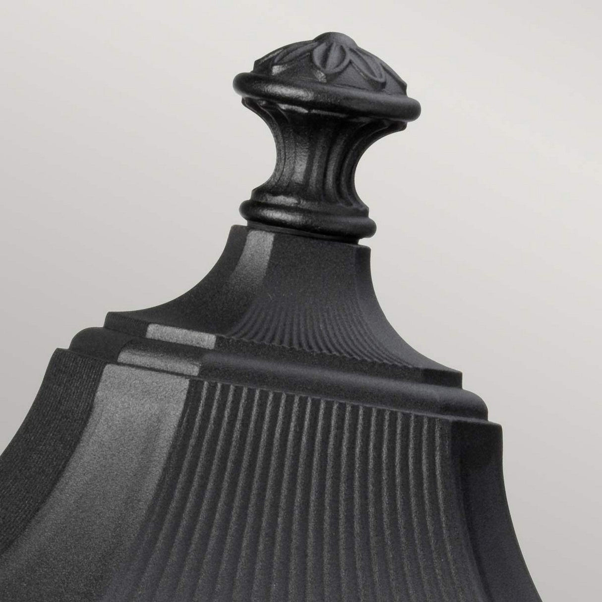 A detailed view of the ornate top of the Cotswold Lane 3 Light Outdoor Large Pedestal in black, showcasing a decorative finial. The design includes vertical grooves and a rounded top adorned with carved leaf patterns, presenting an elegant black finish against a light gray background.