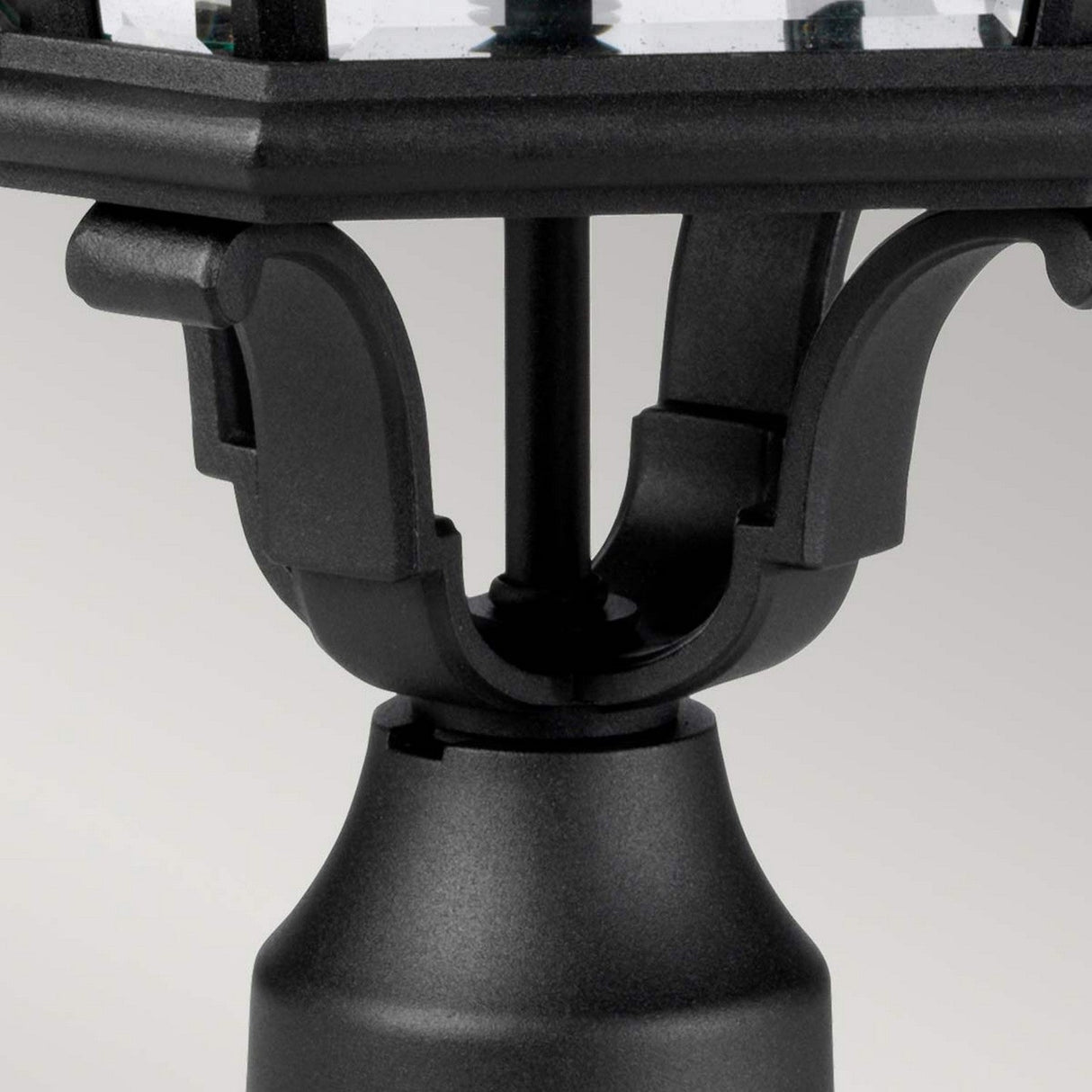 Close-up of the Cotswold Lane 3 Light Outdoor Large Pedestal in a black finish, showcasing a decorative bracket. This design includes a cylindrical base with ornate, curved supports extending upwards. The elegant contrast of the clear glass light fixture is highlighted against the plain light gray background.
