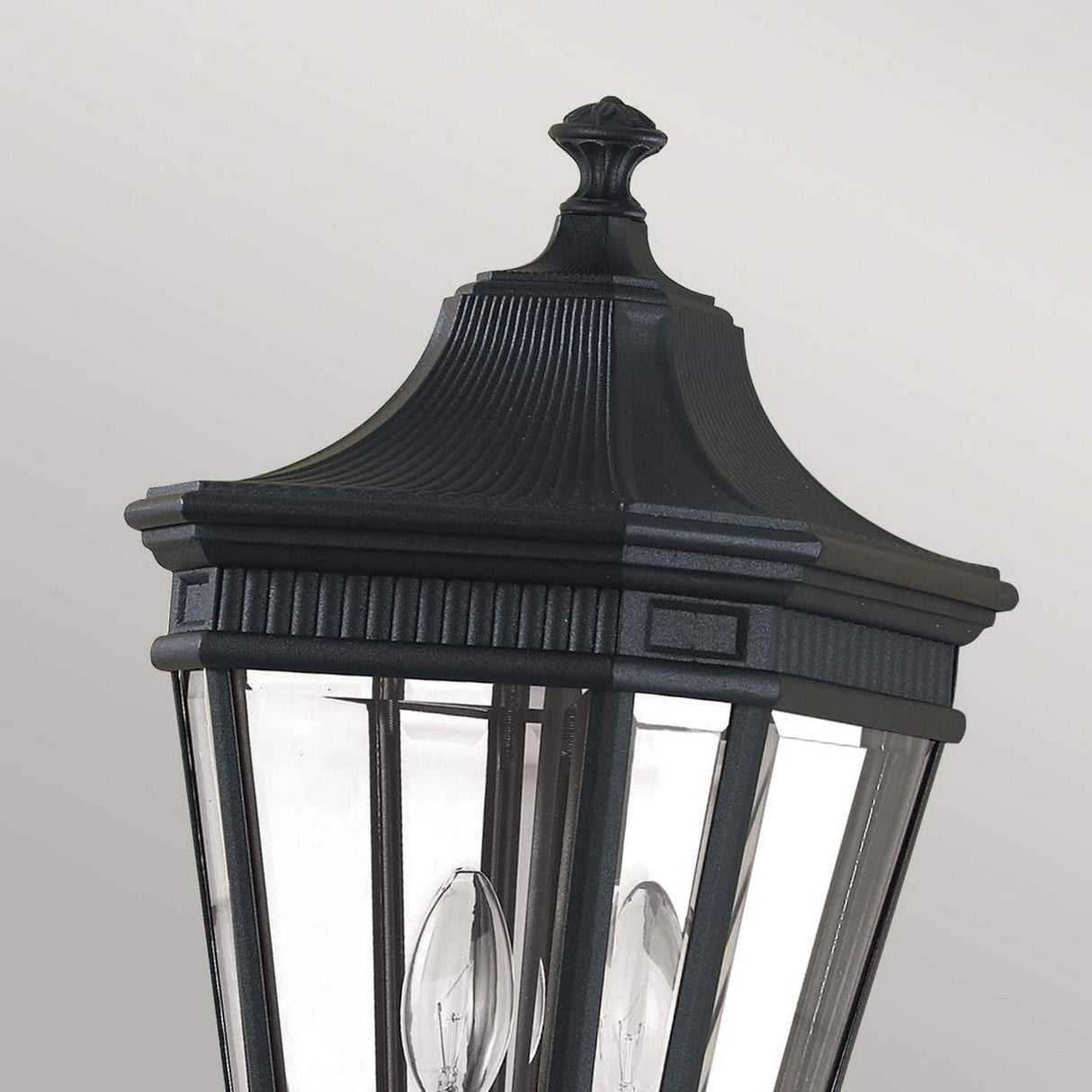 Close-up of the Cotswold Lane 2 Light Outdoor Medium Pillar in black, showcasing its elegant design reminiscent of antique gas lanterns with a decorative, ridged top. The glass panels reveal candle-style light bulbs inside, making it a perfect outdoor lighting fixture against a plain, light gray background.