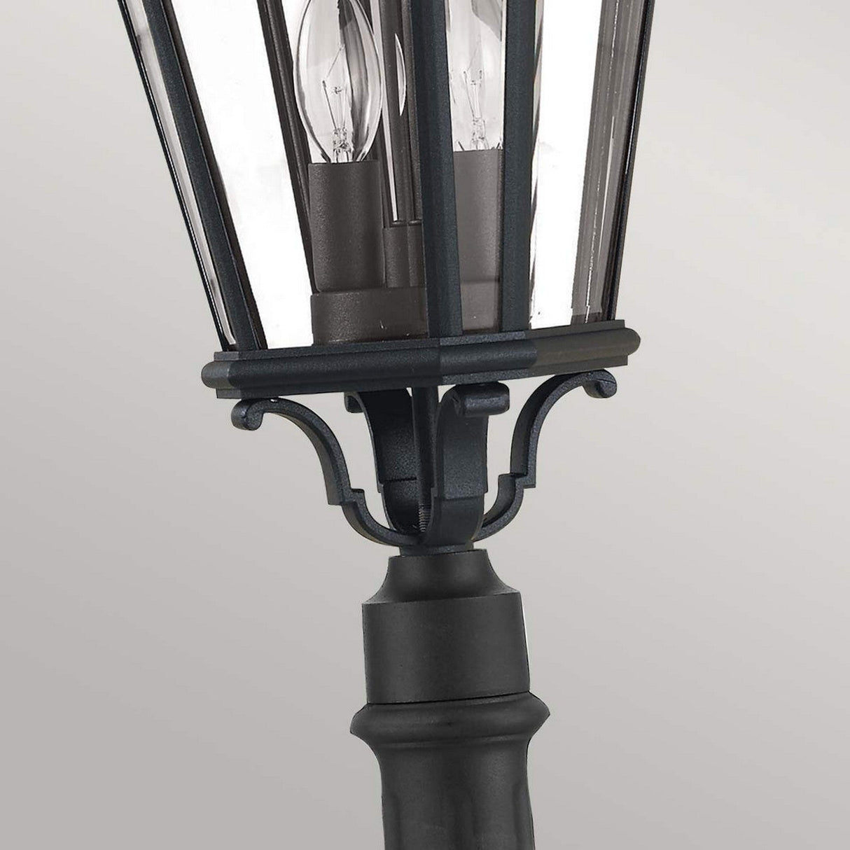 Close-up of the Cotswold Lane 2 Light Outdoor Medium Pillar in black, featuring four glass panels that mimic antique gas lanterns. The fixture displays two visible light bulbs inside, set against a light gray background.