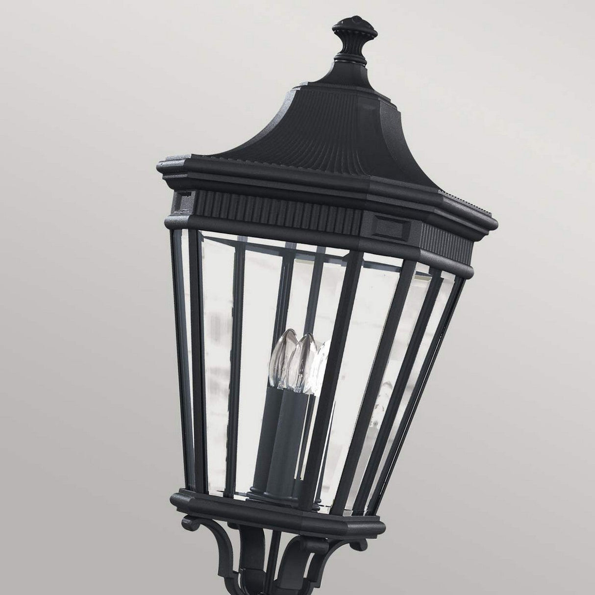 A close-up view captures the elegance of the Cotswold Lane 3 Light Large Post - Black, showcasing its glass panels and three visible bulbs inside. Set against a light gray background, this lantern is reminiscent of classic Cotswold Lane outdoor lighting with its decorative detailing and pointed top.