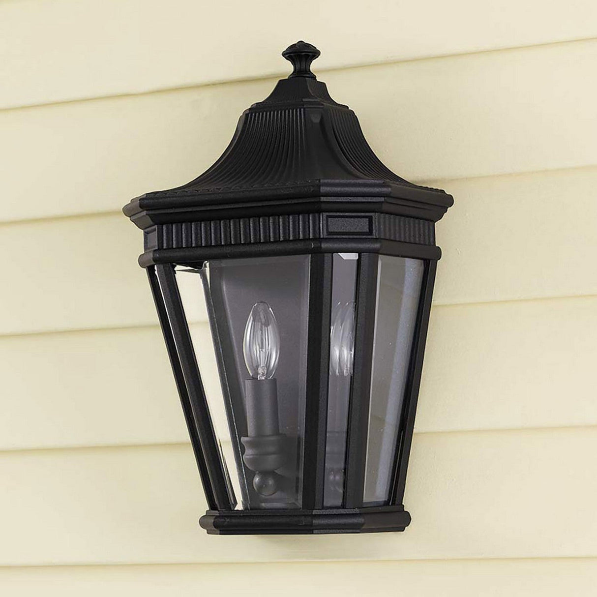 The Cotswold Lane 2 Light Outdoor Wall Lantern - Black is installed on a light-colored siding wall, featuring a traditional design with clear beveled glass. The classic black finish complements the visible candle-style light bulbs elegantly housed inside.