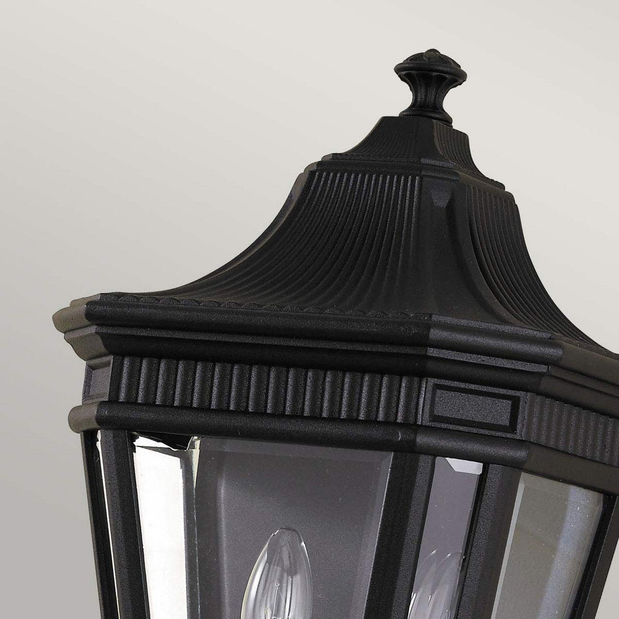 A close-up view of the Cotswold Lane 2 Light Outdoor Wall Lantern in black showcases its sophisticated design with a fluted cap and clear beveled glass panes. This vintage-style outdoor wall light is adorned with intricate details and houses a light bulb inside, set against a soft, neutral background.
