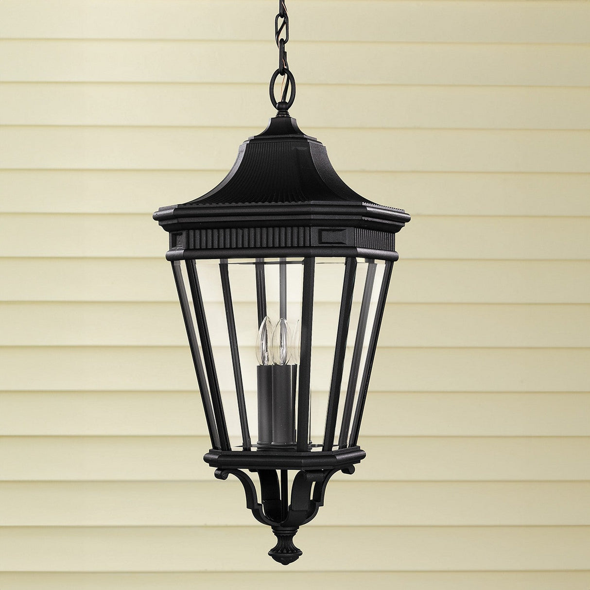 A black Cotswold Lane 3 Light Outdoor Large Chain Pendant exuding vintage charm hangs from a chain against a beige background with horizontal siding. This outdoor lighting piece features clear glass panels and two visible candle-shaped bulbs inside.