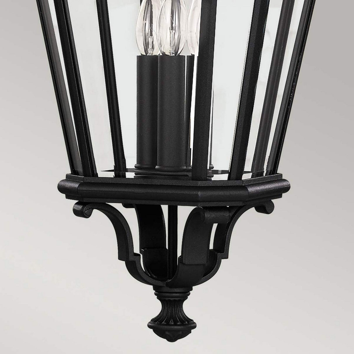 A close-up of the Cotswold Lane 3 Light Outdoor Large Chain Pendant in black showcases its vintage charm, featuring vertical glass panels and three candle-style light bulbs. With ornate detailing against a plain gray background, it embodies elegant outdoor lighting solutions.