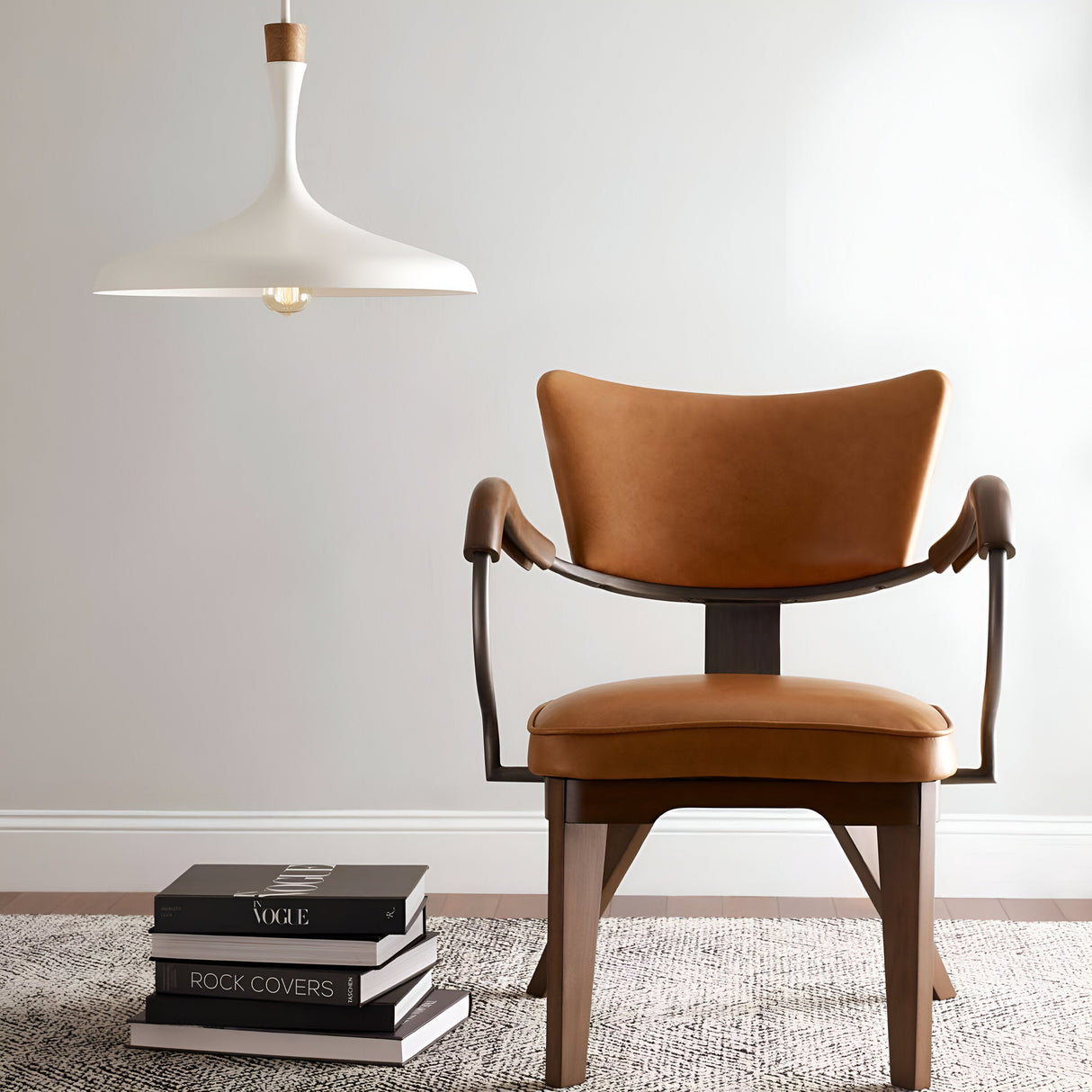 A modern living room scene features a tan leather chair on a gray rug, with a stack of books nearby on the floor. Above, the Darwin 1 Light Pendant in Matte White hangs, its adjustable height allowing it to cast an ambient glow that enhances the soft gray walls and complements the urban style of the minimalist decor.
