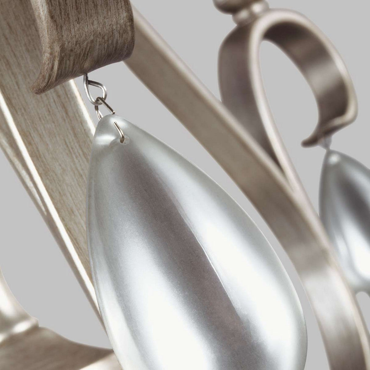 A close-up of the DeWitt 5 Light Chandelier in silver, featuring teardrop-shaped metal ornaments hanging from a decorative, curving metallic design, reminiscent of French Provincial style, with a light wood finish. The soft gray background enhances its minimalist and elegant aesthetic.