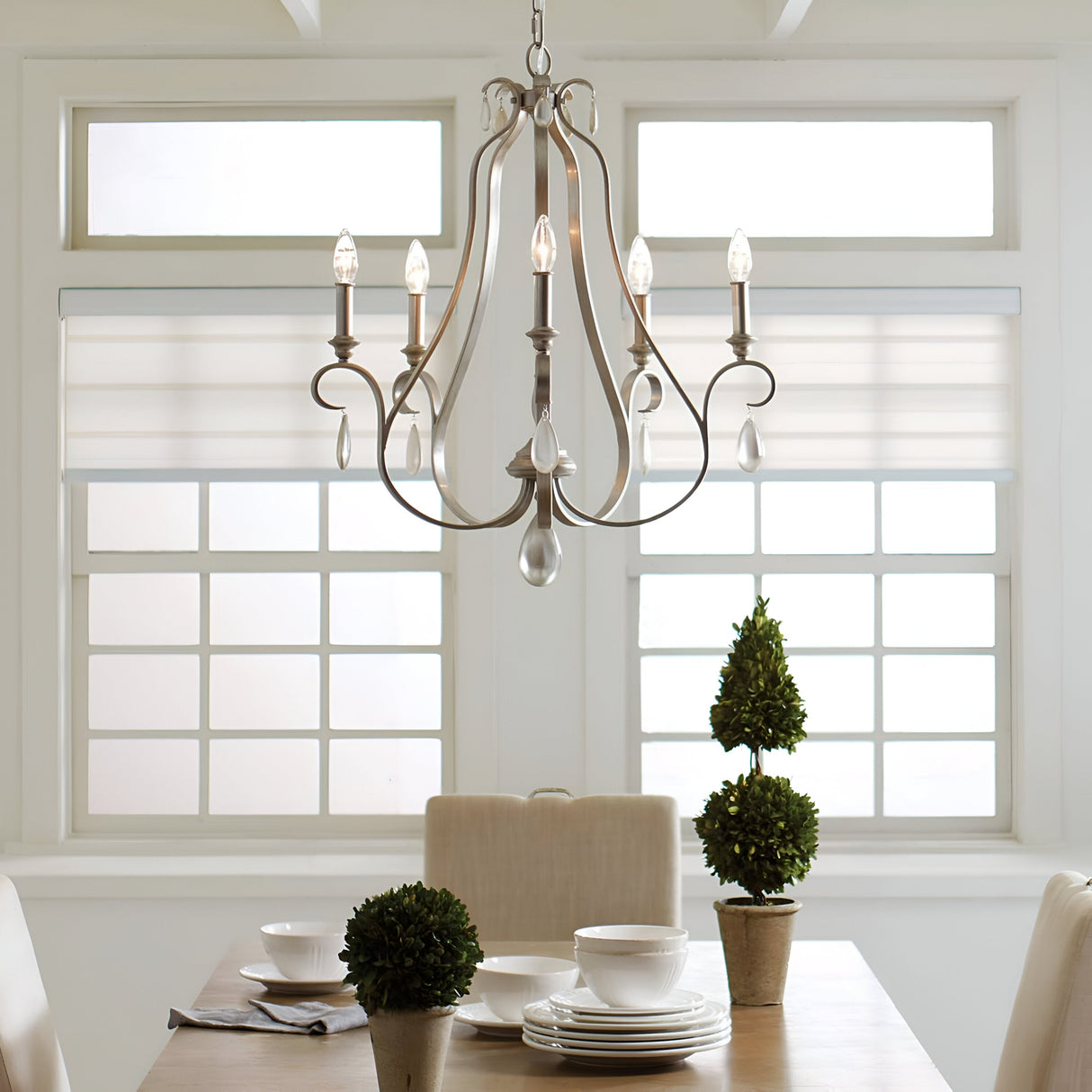In a dining room showcasing a wooden table set for four, complete with white dishware and small potted topiary plants, the graceful elegance of the DeWitt 5 Light Chandelier in silver adorned with teardrop crystals enhances the ambiance. The French Provincial style is further embraced by large windows fitted with white blinds that brighten the space.