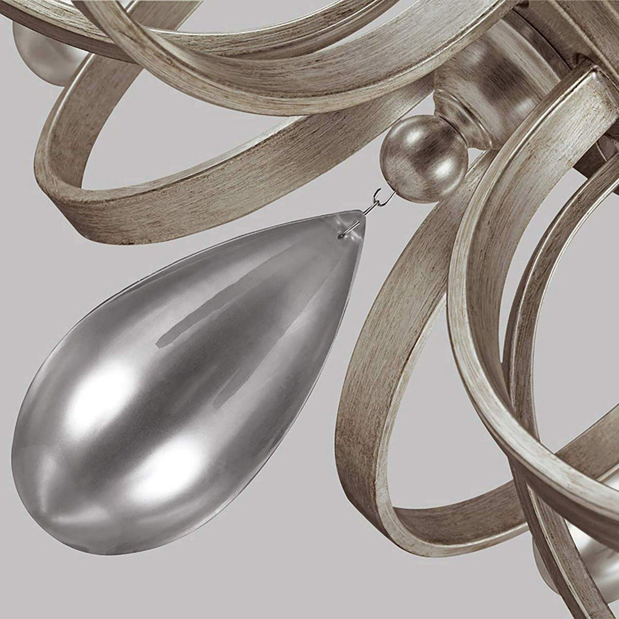 A close-up of the DeWitt 9 Light Chandelier - Silver showcases intertwined metallic ribbons and glossy, teardrop crystals. Its combination of smooth and textured surfaces delivers an elegant, modern look with a touch of French Provincial design.