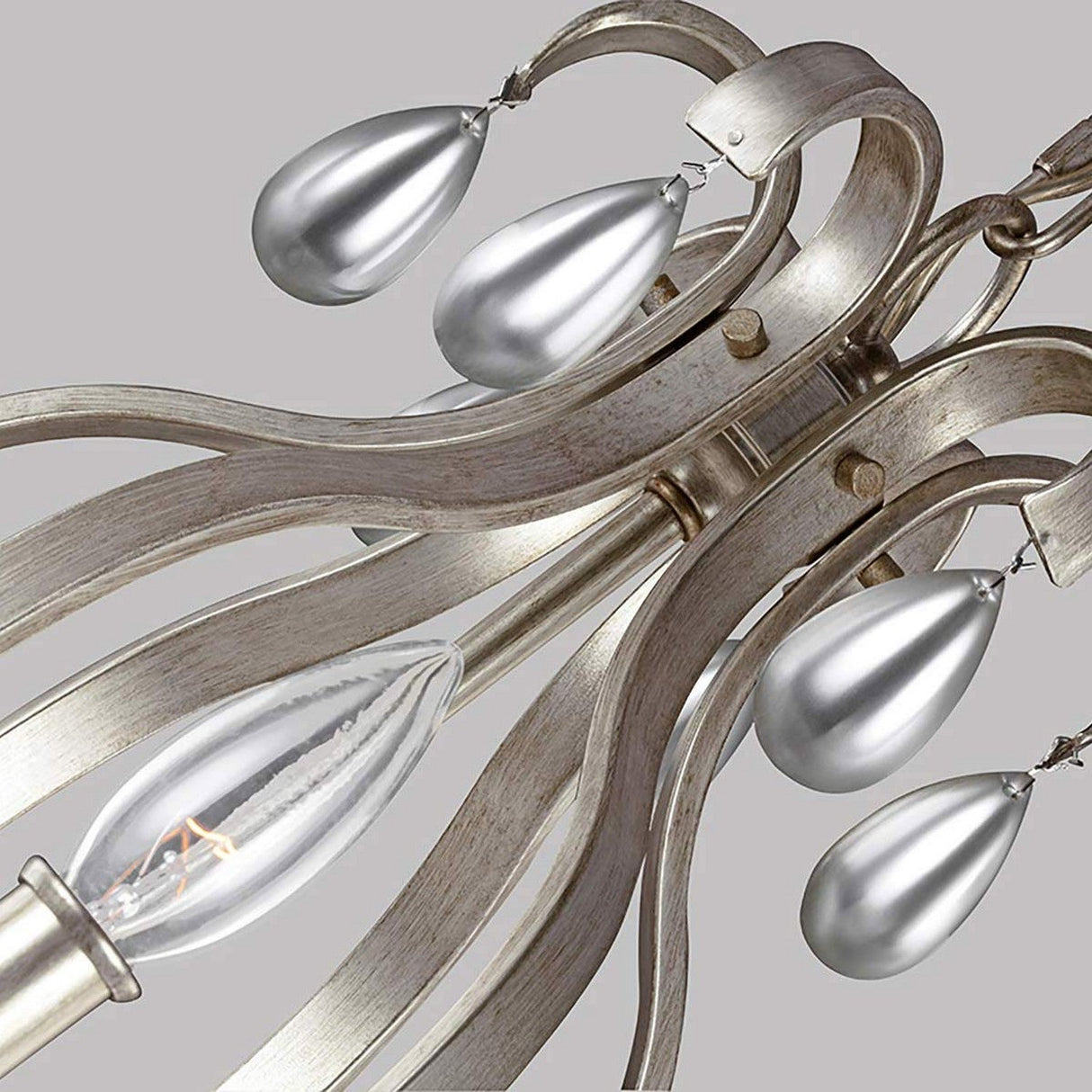 Close-up of the DeWitt 9 Light Chandelier - Silver, featuring elegantly curving metallic arms and teardrop-shaped silver ornaments. An exposed bulb enhances the modern appeal of this intricate, French Provincial design against a neutral backdrop.