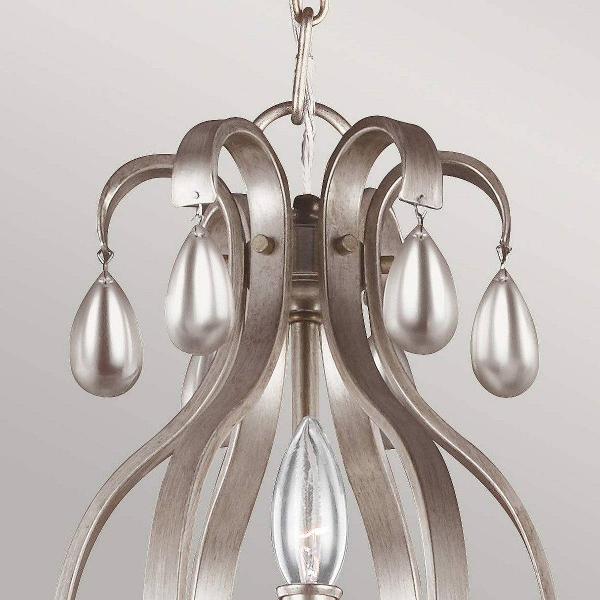 A close-up of the sophisticated DeWitt 9 Light Chandelier - Silver highlights its gracefully curved metallic arms adorned with teardrop crystals. A single clear bulb softly illuminates, evoking a touch of French Provincial style against a neutral background.