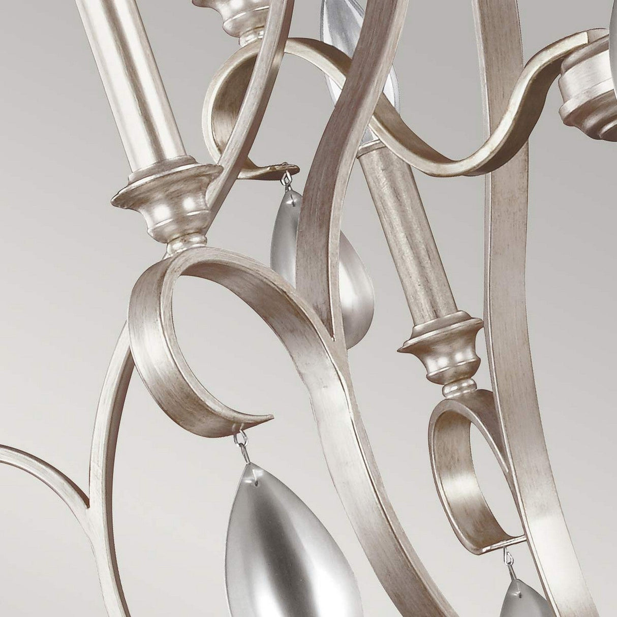 A close-up of the DeWitt 9 Light Chandelier - Silver showcases its swirling metallic arms and teardrop crystals that delicately hang, reflecting light. The chandelier's French Provincial design and exquisite craftsmanship are highlighted against a neutral gray background.