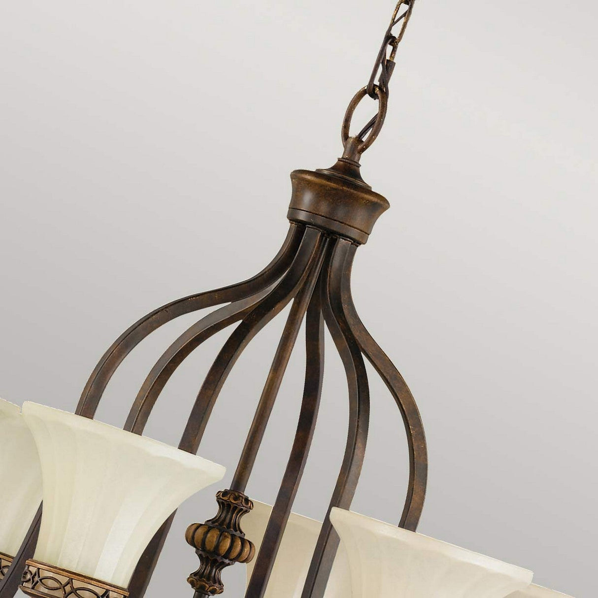 The Drawing Room 6 Light Chandelier – Walnut showcases a close-up view of an elegant Edwardian style design, featuring a bronze-colored, ornate metal frame with several frosted glass shades. This sophisticated fixture is suspended from the ceiling by a metal chain and highlights a classic aesthetic with a subtle walnut finish.