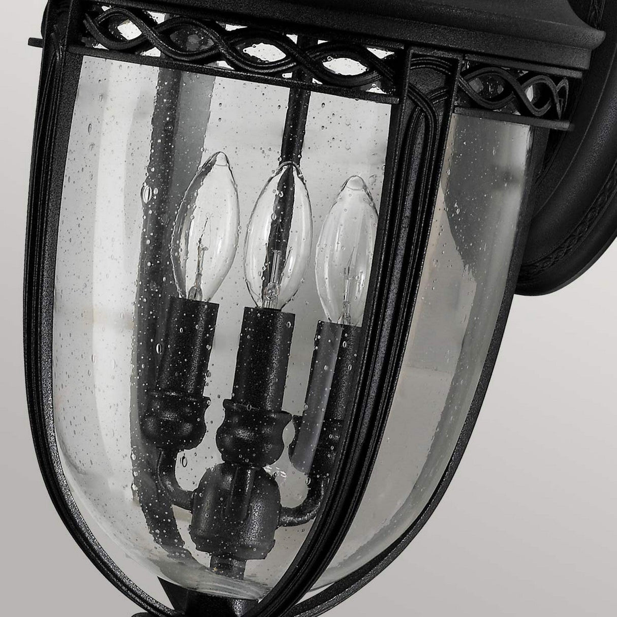 A close-up of the English Bridle 3 Light Outdoor Large Wall Lantern - Black highlights its clear seeded glass and black metal frame. The lantern, with its trio of candle-shaped bulbs, is adorned with raindrops, hinting at a recent or ongoing rain.