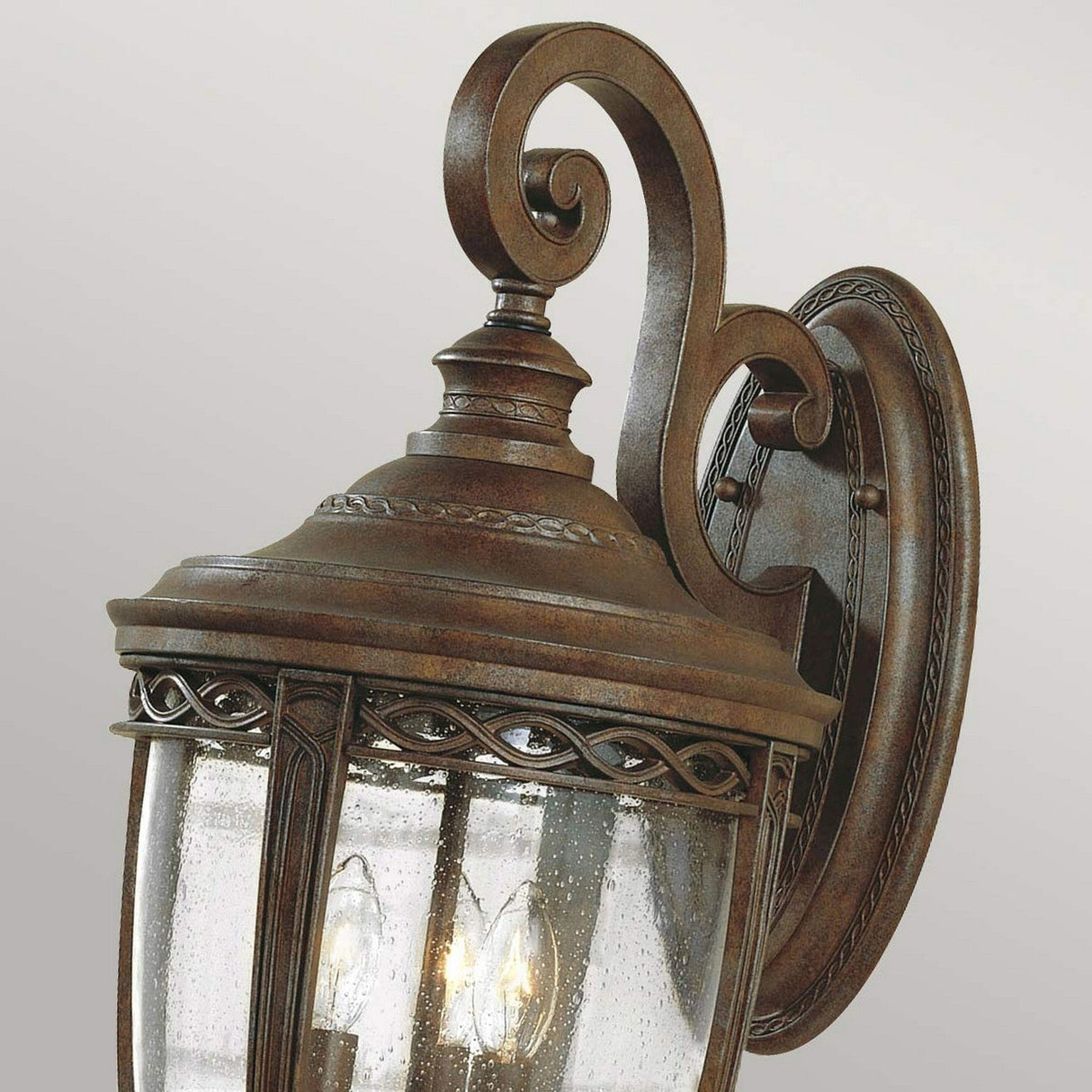 A detailed view of the English Bridle 3 Light Outdoor Large Wall Lantern - Bronze highlights its intricate scrollwork and seeded glass panels, set against a plain background. Crafted for wall mounting, this lantern exudes the elegance of outdoor lighting with its decorative detailing.