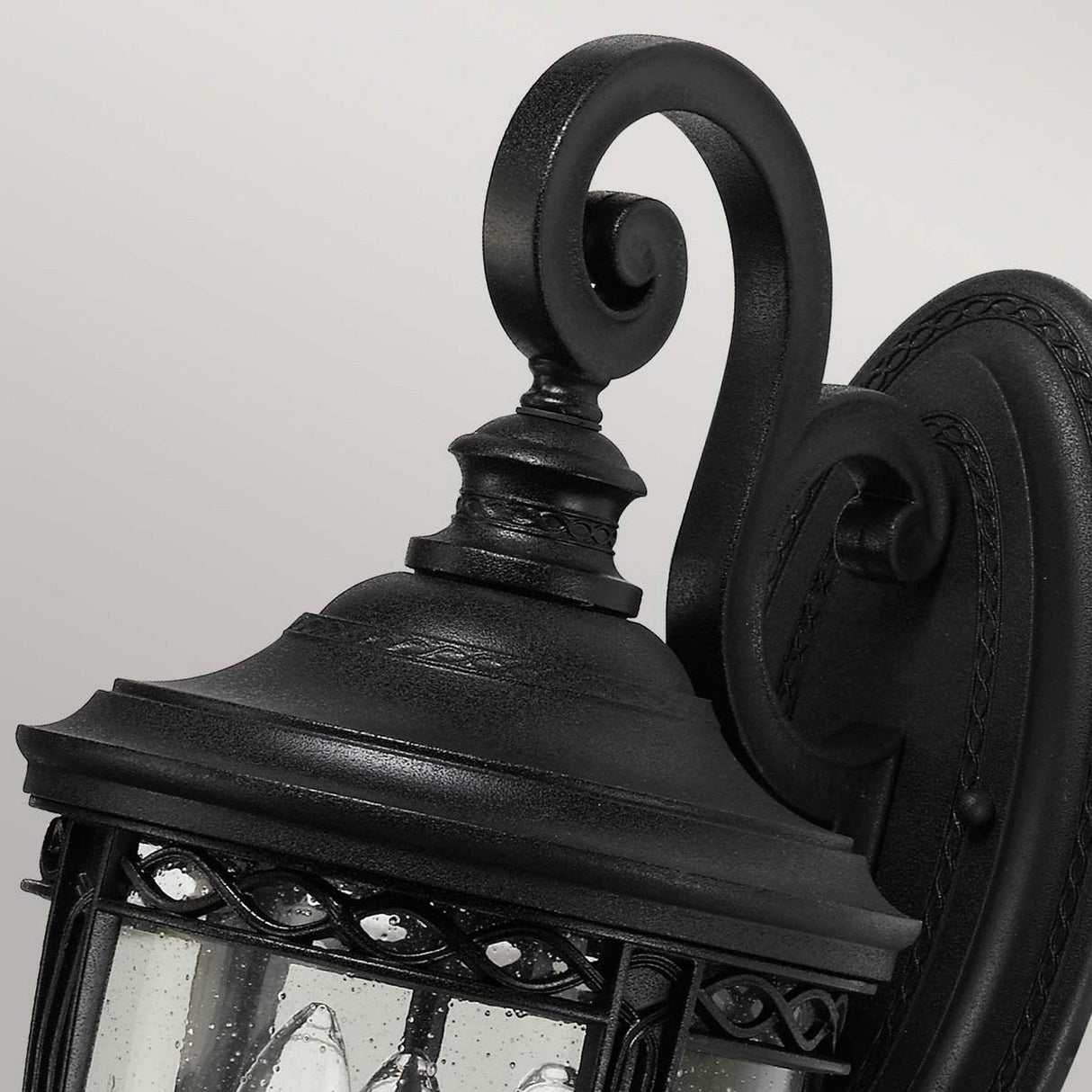 Close-up of the English Bridle 3 Light Outdoor Medium Wall Lantern in black, featuring a curved and ornate wrought iron design. The lantern showcases clear seeded glass panels that reveal the internal light bulbs, standing out beautifully against the plain, light gray background.