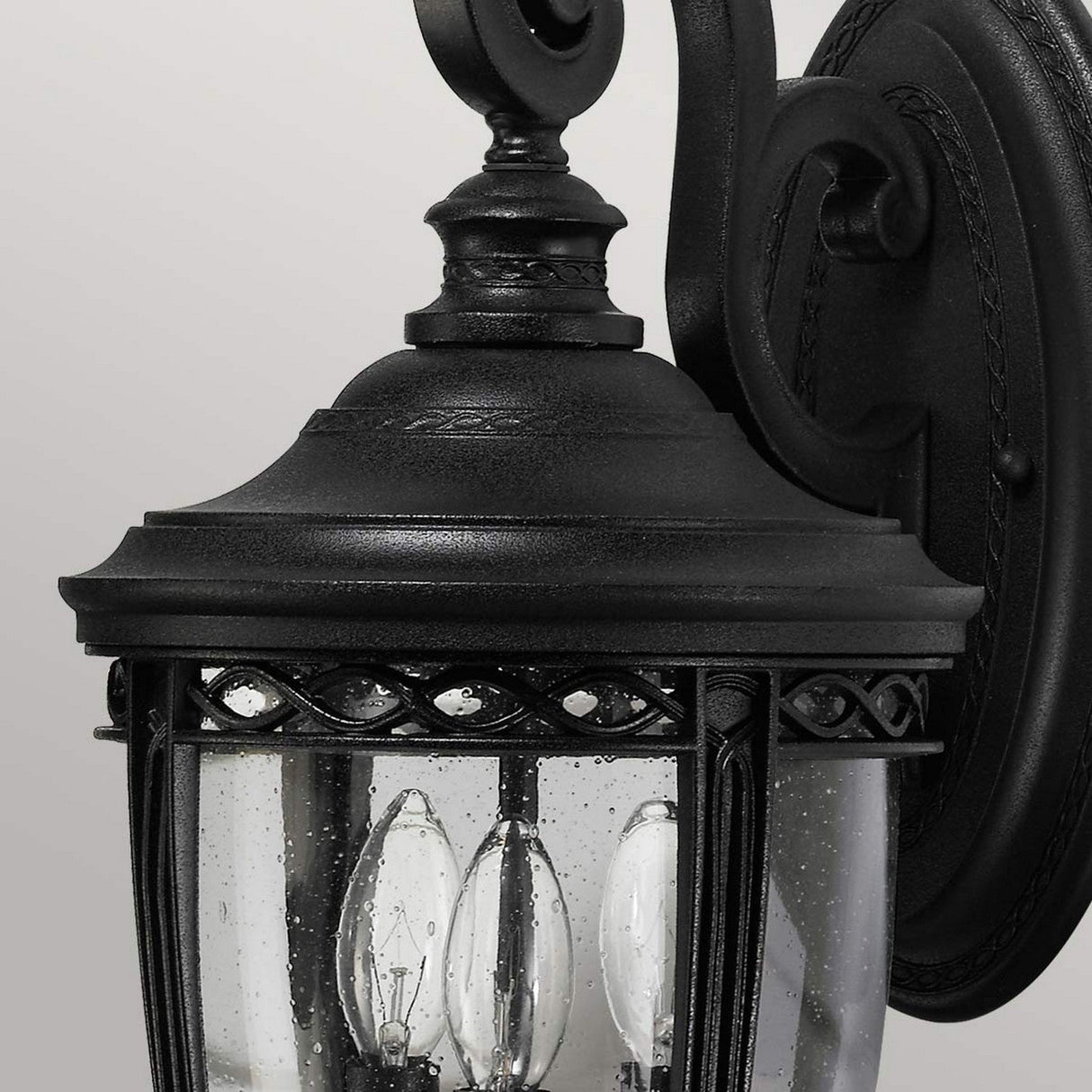 Close-up of the English Bridle 3 Light Outdoor Medium Wall Lantern - Black, showcasing its black metal frame and clear seeded glass panels. Inside, three candle-shaped bulbs emit a warm glow. This vintage-style outdoor wall lantern features ornate detailing and is mounted on a gracefully curved bracket.