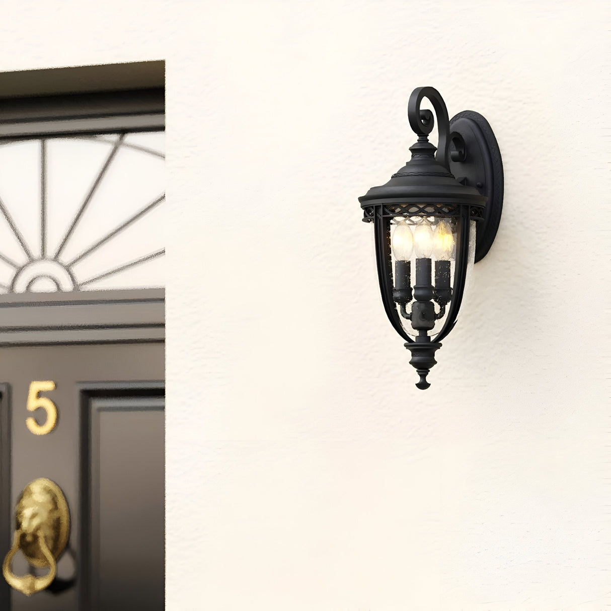 The English Bridle 3 Light Outdoor Medium Wall Lantern in black, featuring clear seeded glass panels, beautifully lights up the cream-colored wall. Two bulbs emit a warm glow beside a dark door equipped with a brass knocker and the house number 5.
