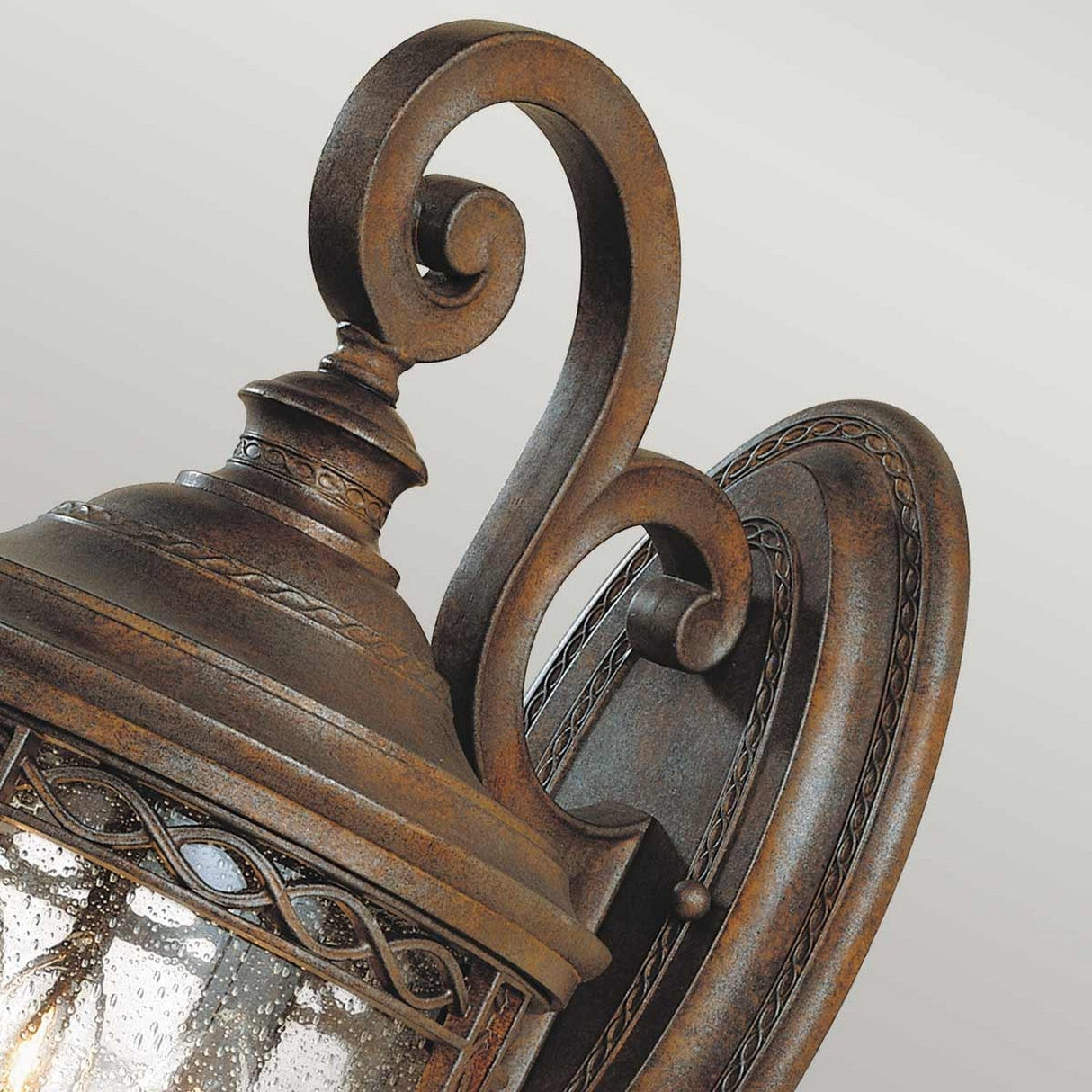 Close-up of the English Bridle 3 Light Outdoor Medium Wall Lantern in Bronze, featuring ornate vintage-style design with intricate scrollwork and a clear seeded glass cover, elegantly mounted on a decorative oval backplate.
